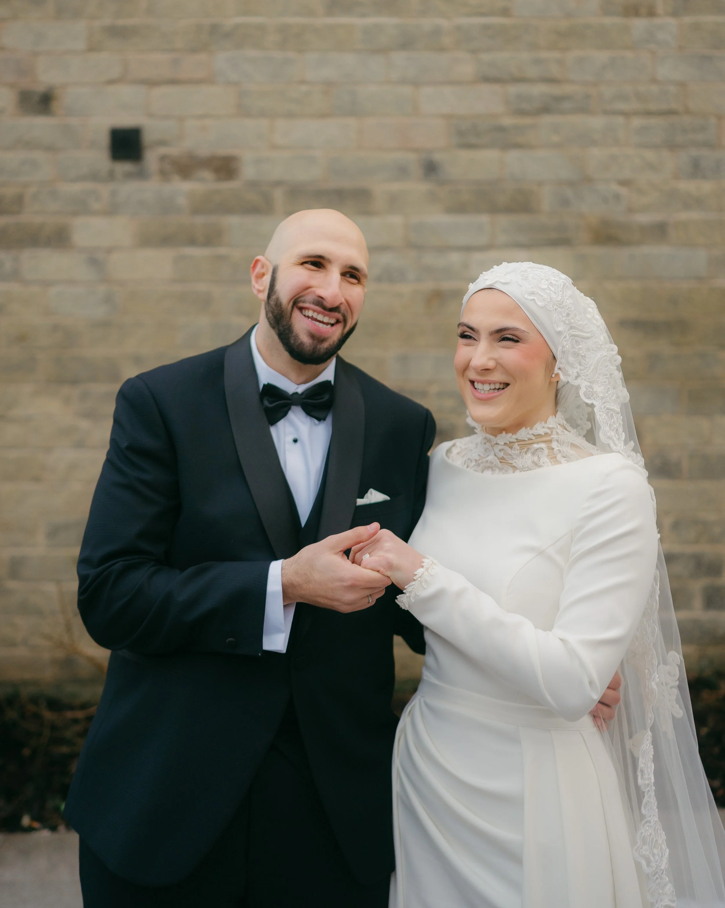 A bride and groom smiling and holding hands against a brick wall.