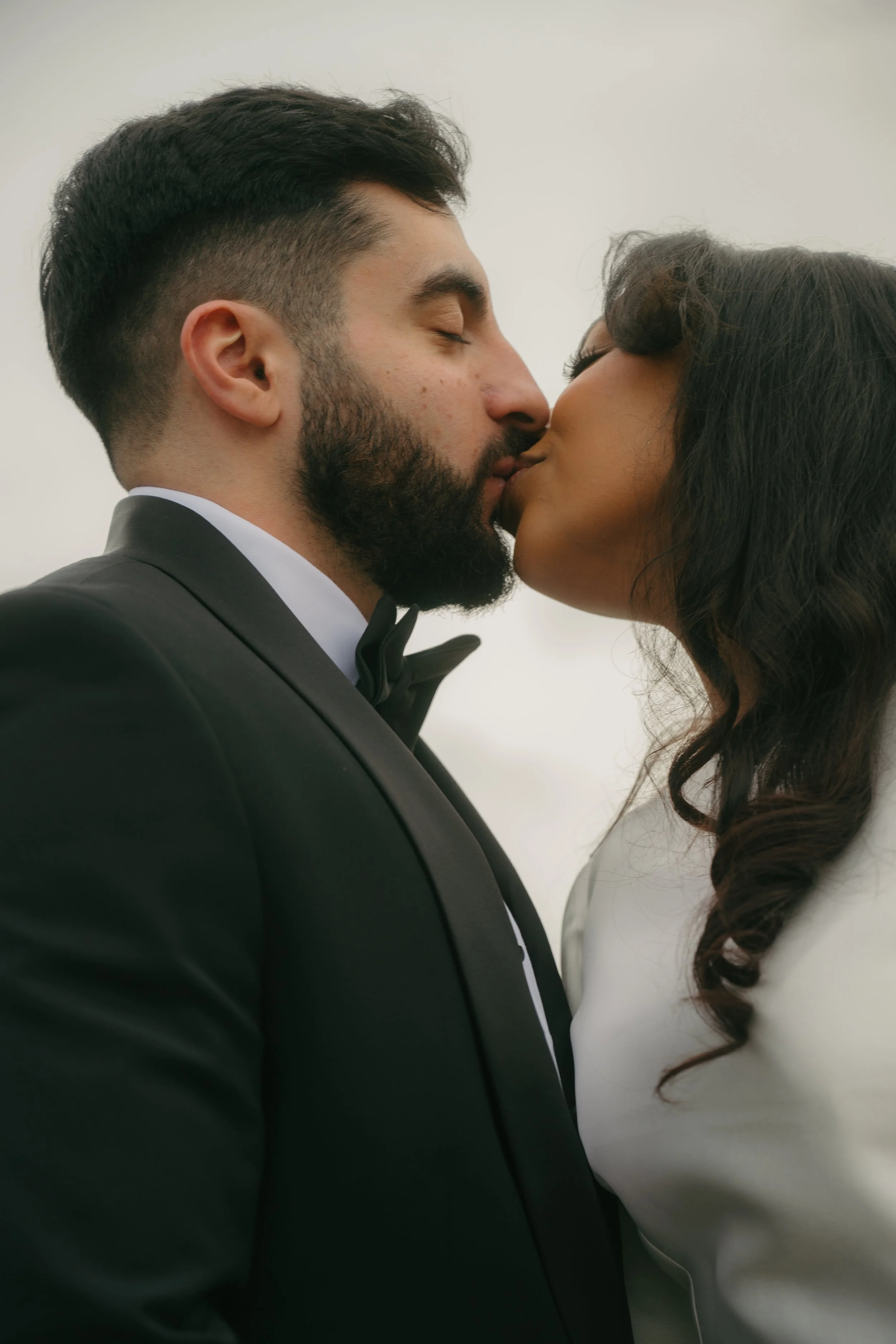 A man in a black tuxedo and a woman in a white dress are kissing, outdoor, wedding or formal event.