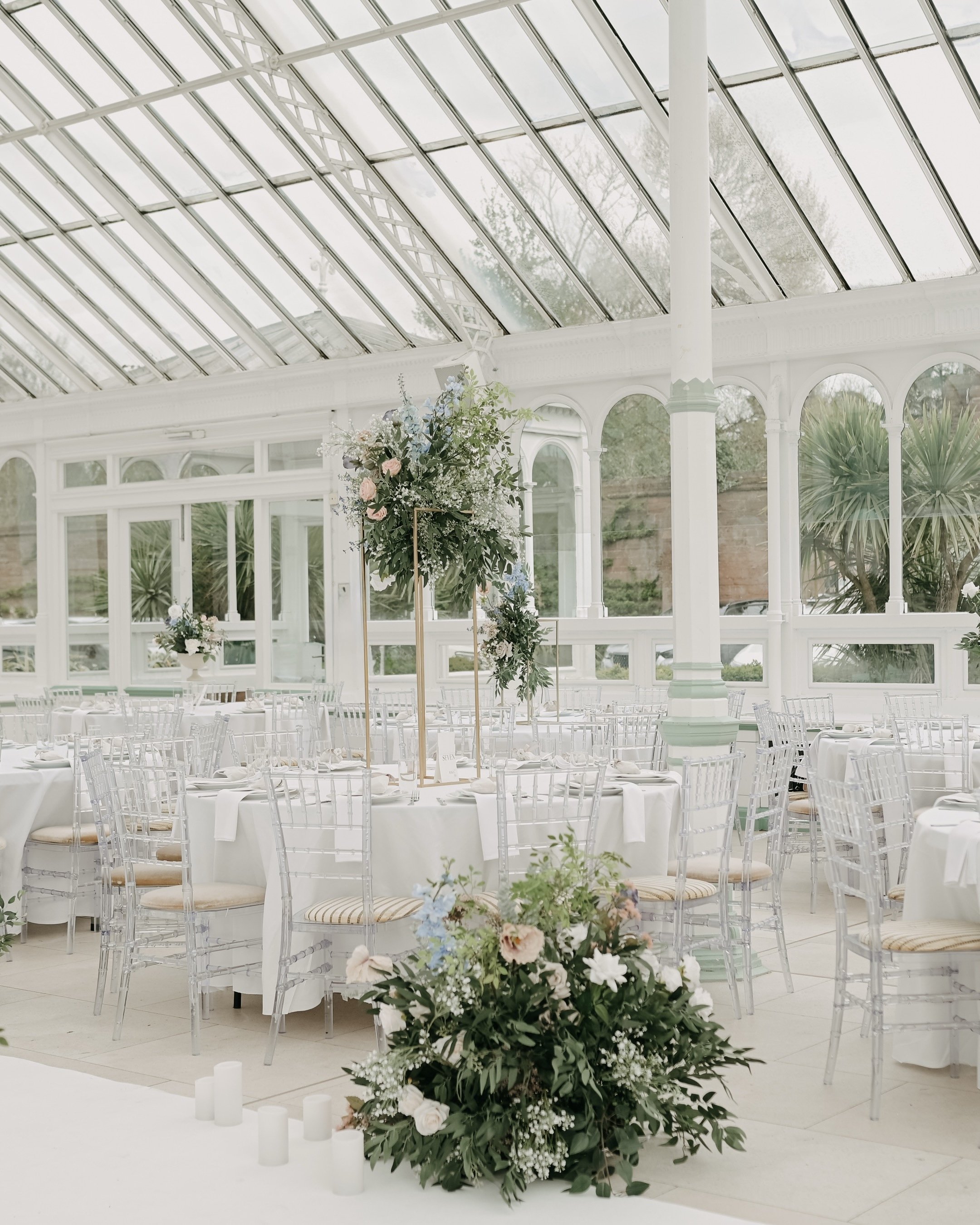 Elegant wedding reception setup with round tables, white tablecloths, floral centerpieces, and transparent chairs inside a glass conservatory.