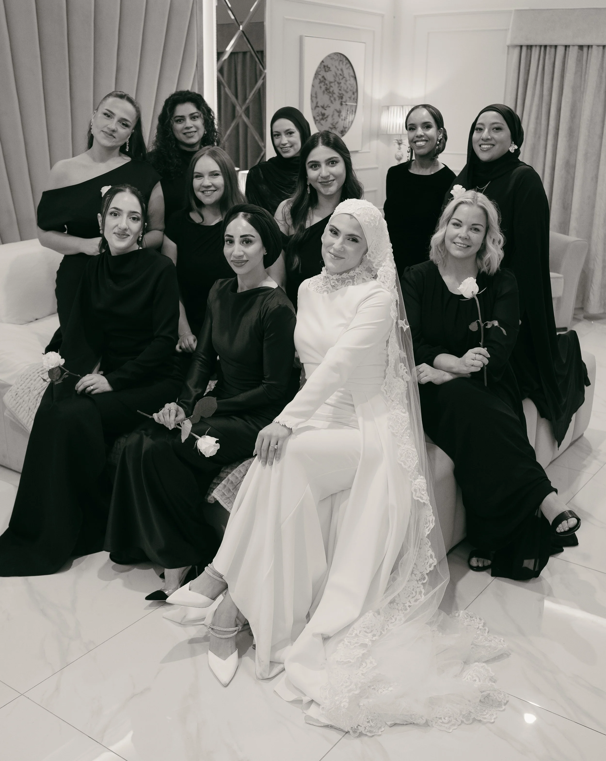 A group of women, including a bride in a white dress and hijab, sitting and standing in a hotel lobby with curtains, mirror, and lamp, all dressed in black or dark clothing, some holding white roses.