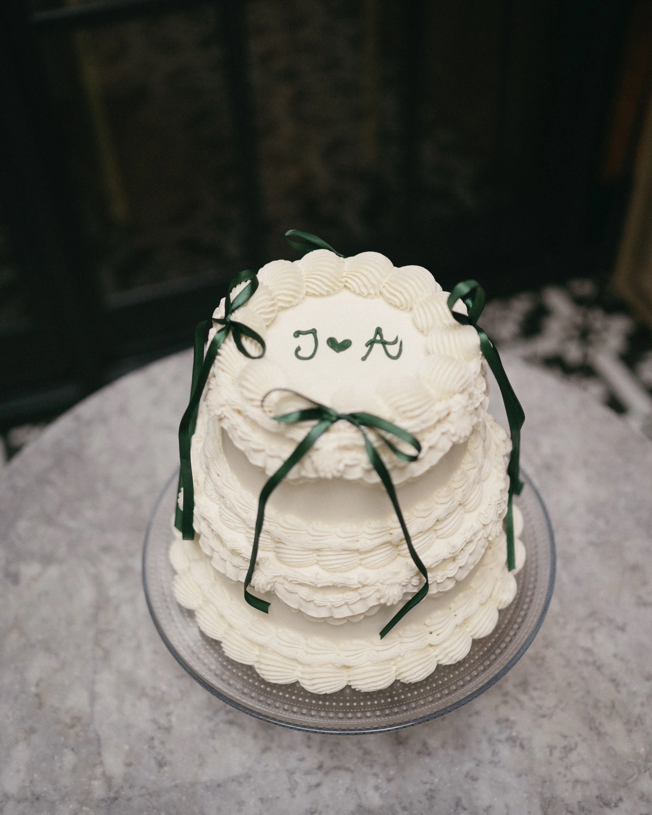 A three-tier white wedding cake decorated with green ribbons, with the top tier inscribed with initials 'J' and 'A' and a heart in between, on a glass cake stand.