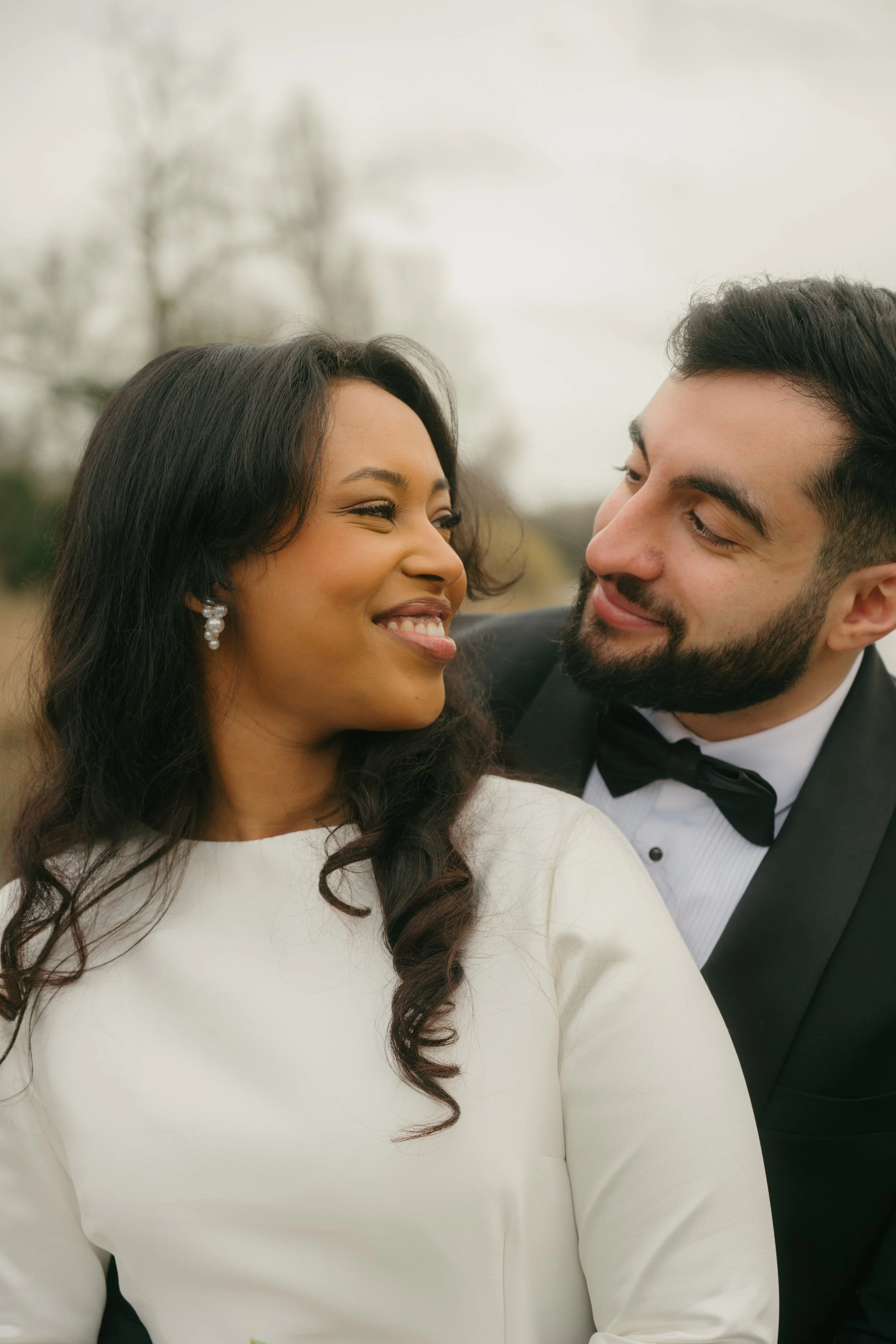 A joyful interracial couple, dressed in wedding attire, smiling and looking into each other's eyes outdoors on a cloudy day.