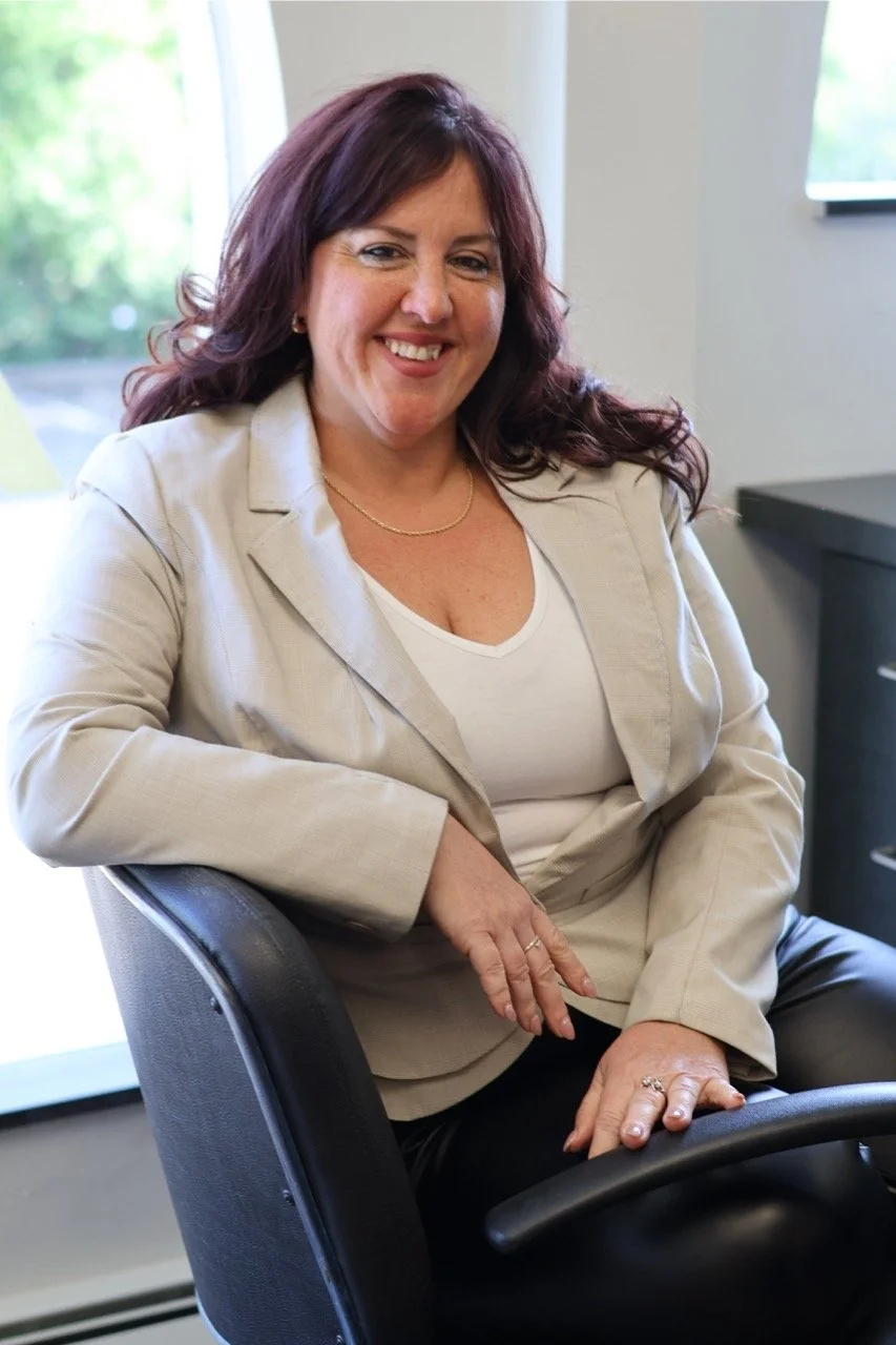 A woman with dark red hair smiling and sitting in an office chair, wearing a beige blazer over a white top, with a window and light-colored wall in the background.