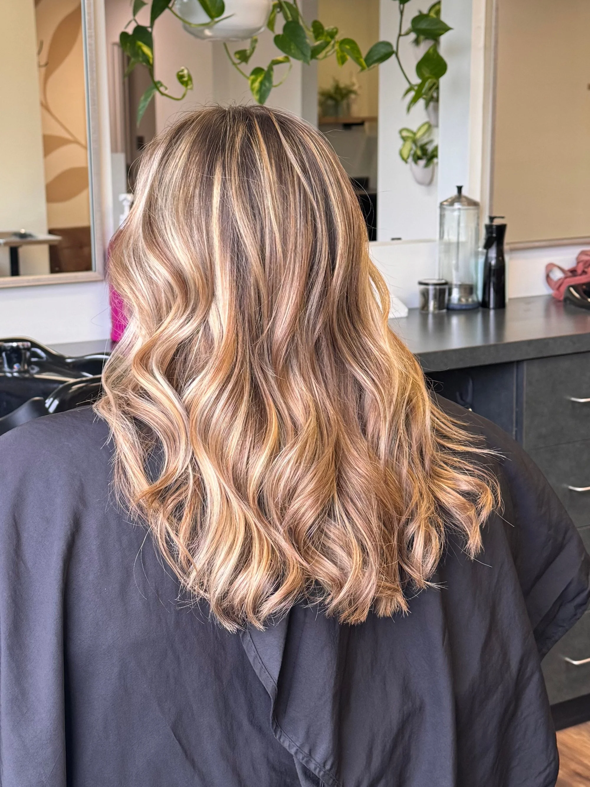 Back view of a person with long, wavy hair showcasing blonde highlights, seated in a salon chair.