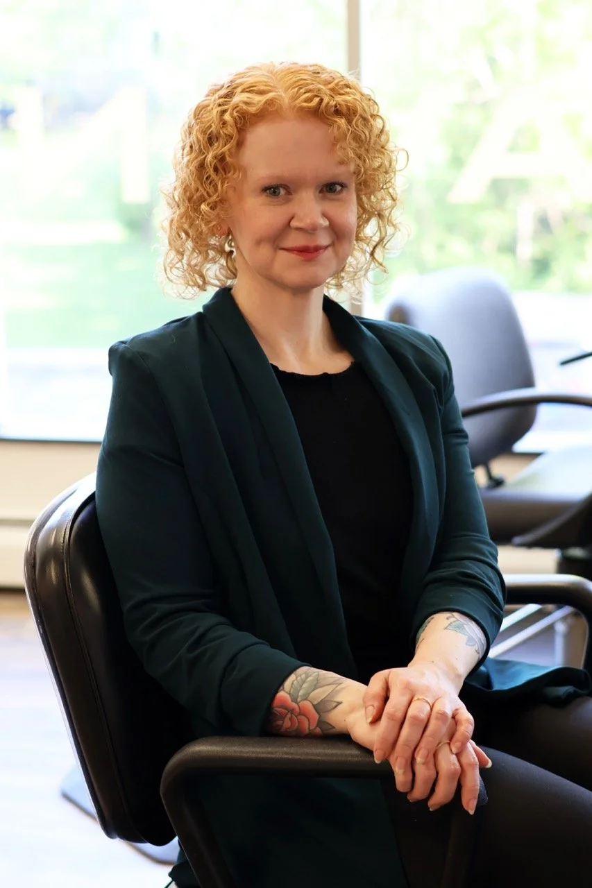 A woman with curly red hair sitting in an office chair, wearing a black top and dark blazer, with tattoos on her forearms, in front of a large window showing green outdoor scenery.