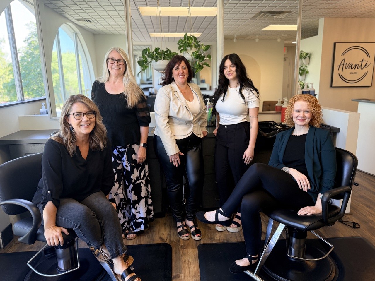 Five women in a hair salon posing for a photo, standing or sitting near salon chairs and mirrors, with a sign reading 'Avanté SALON' visible in the background.
