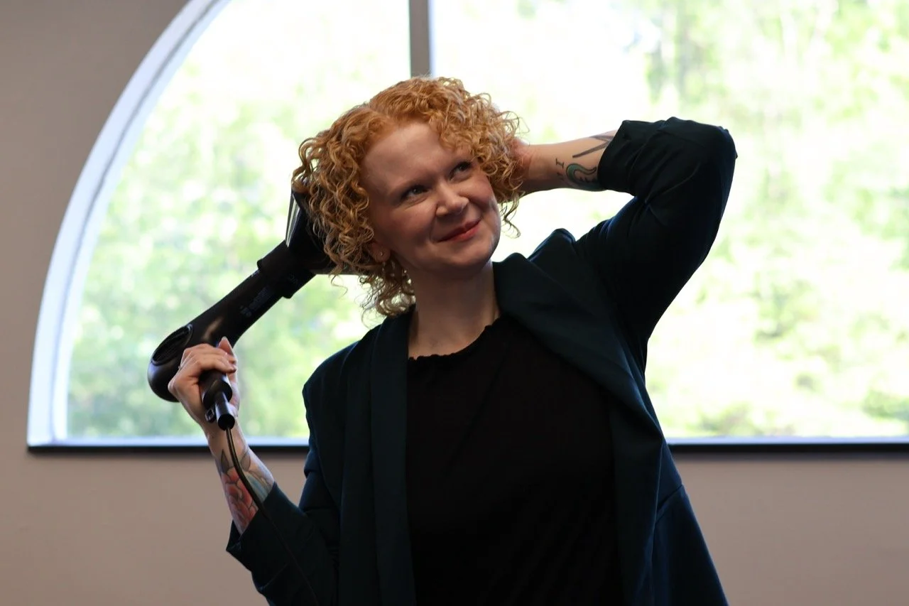 Woman with curly red hair holding a blow dryer on her shoulder and smiling indoors with a large window behind her.
