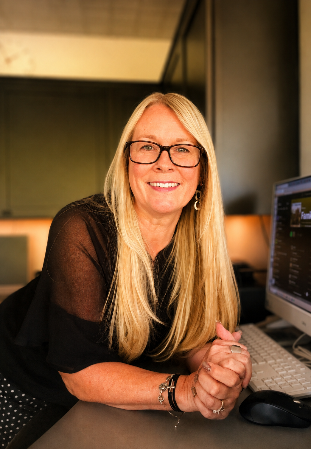 A woman with long blonde hair, glasses, and wearing earrings, sitting at a desk in front of a computer, smiling at the camera.