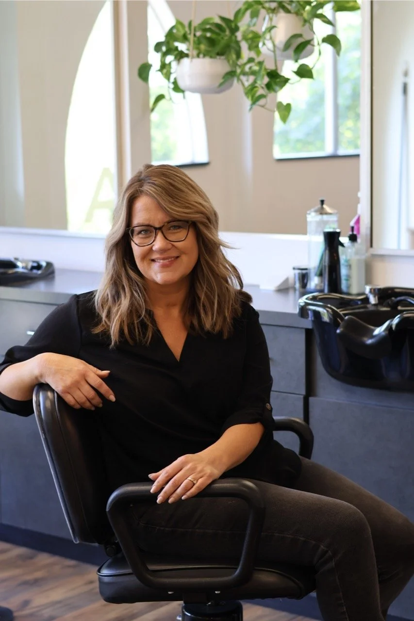 A woman with shoulder-length brown hair, glasses, and a black shirt, sitting in a salon chair inside a hair salon with a large mirror, hair styling tools, and a hanging plant in the background.
