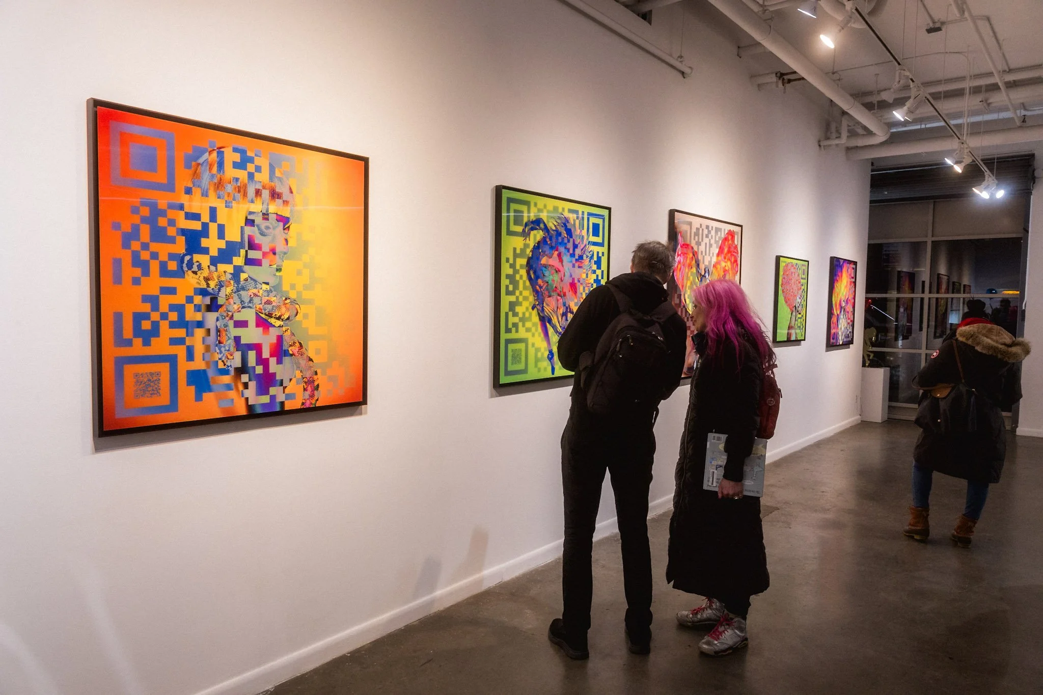 Deux personnes discutent devant une série d'œuvres d'art numérique dans une galerie. Les tableaux comportent des motifs de codes QR avec des images colorées de portraits et d'abstractions. Un autre visiteur marche dans la galerie.