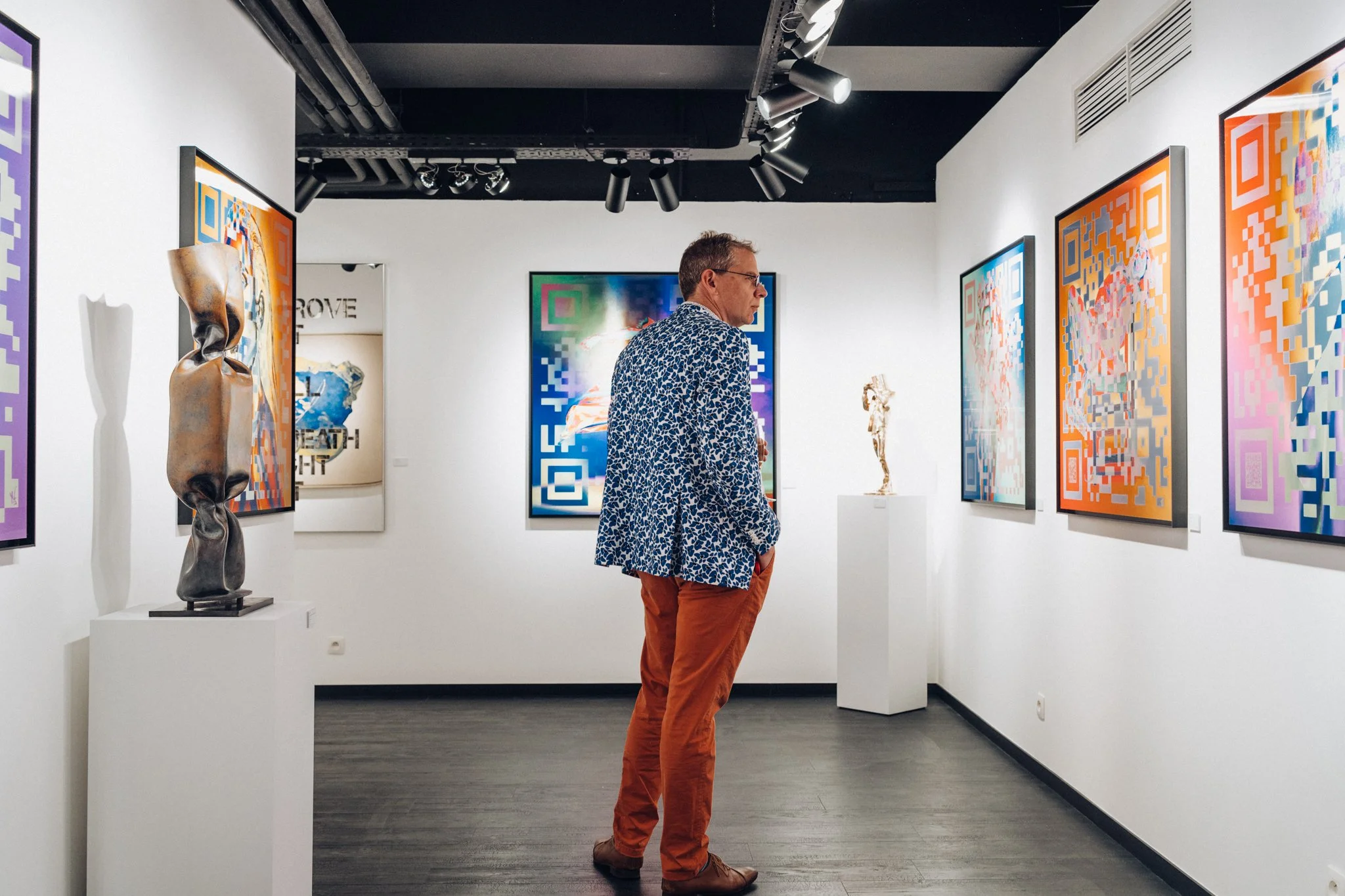 Un homme regarde une œuvre d'art dans une galerie d'art moderne avec plusieurs tableaux colorés et sculptures sur des socles blancs.