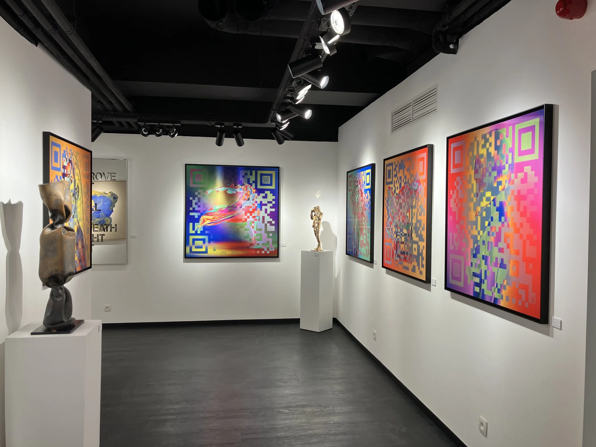 Une galerie d'art moderne avec plusieurs œuvres colorées, peintures avec motifs QR code, sculptures abstraites en bronze, murs blancs, plafond noir avec éclairage directionnel.