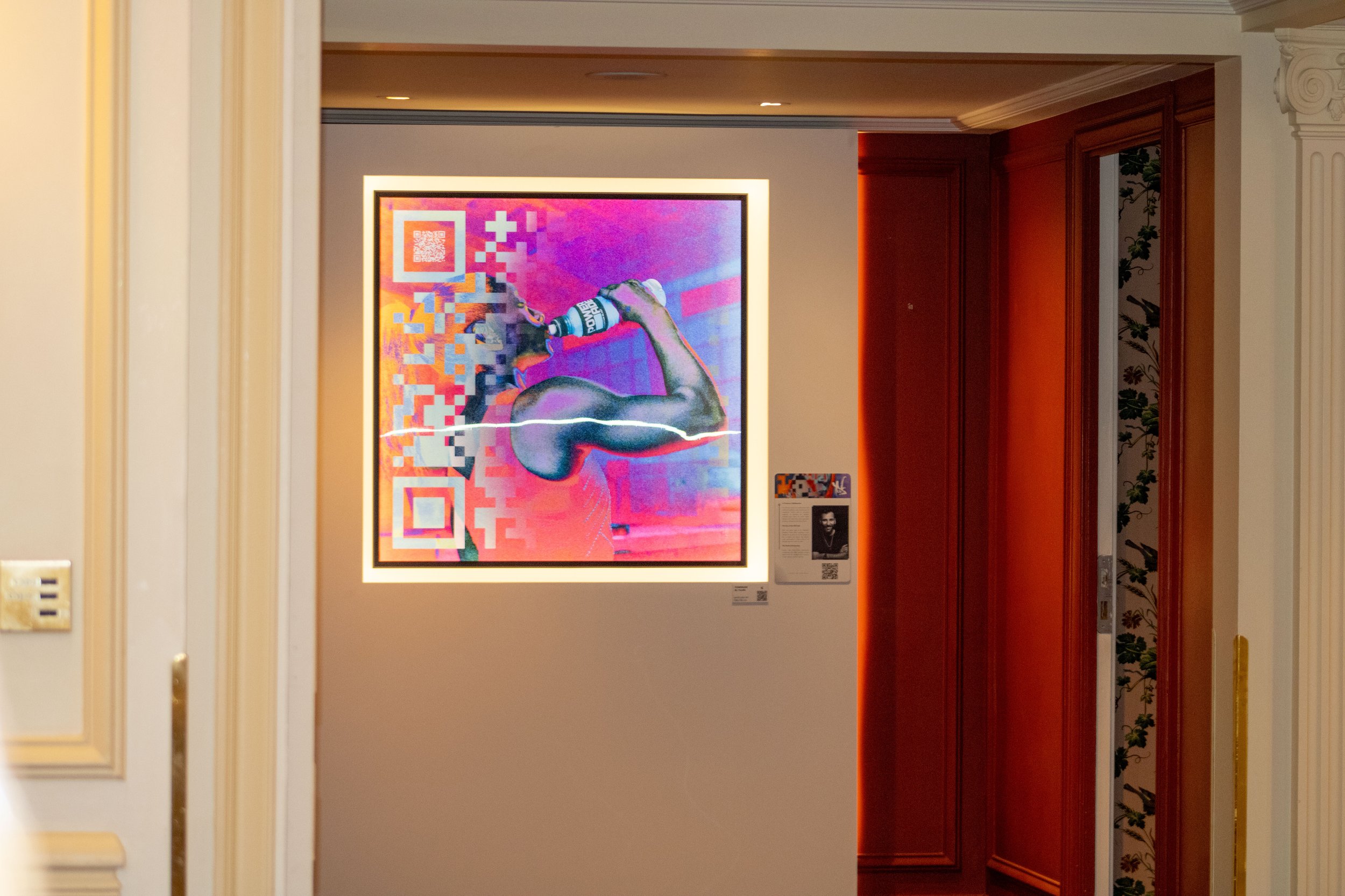 Une œuvre d'art numérique représentant un bras musclé tenant une bouteille, avec un arrière-plan coloré et des motifs en pixels, encadrée et exposée dans une galerie.