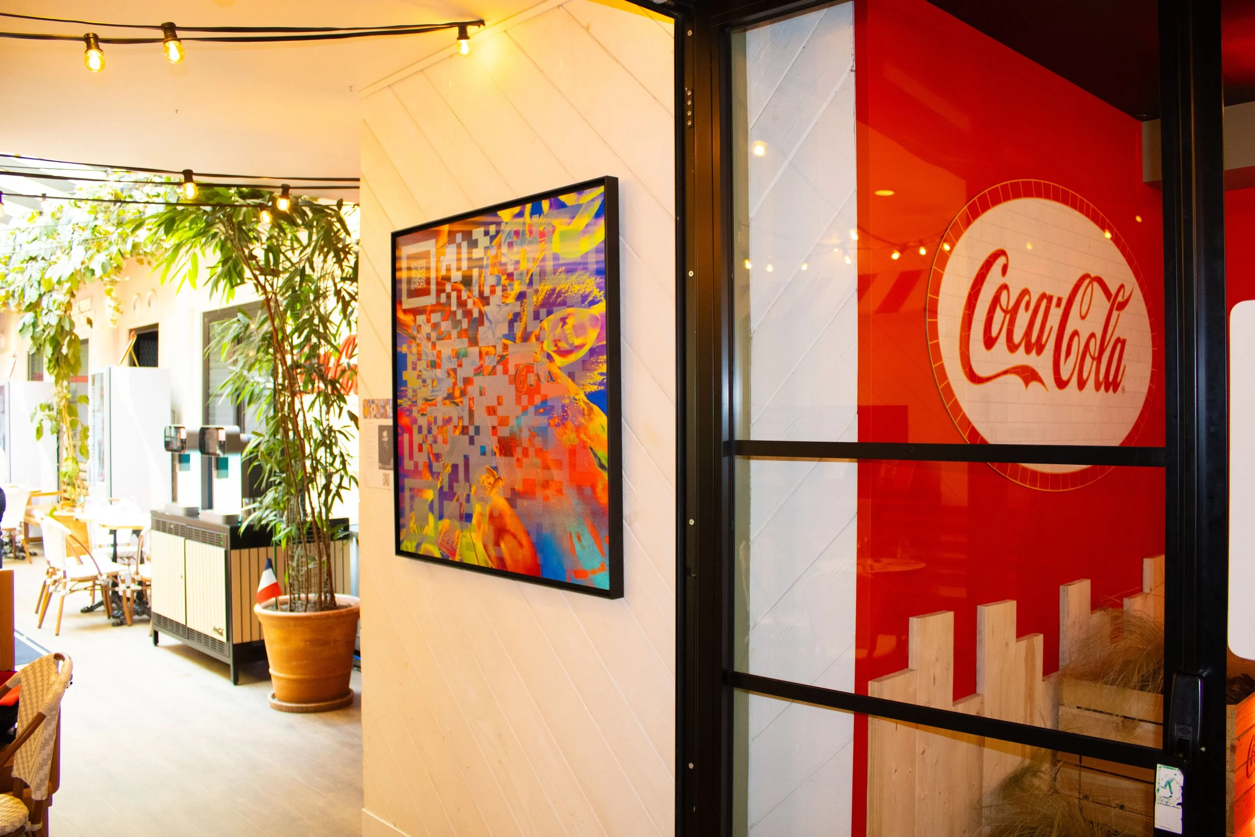 Intérieur d'un restaurant ou café avec décor coloré, tableau abstrait sur un mur blanc, plantes vertes, et une porte en verre avec le logo Coca-Cola en rouge.