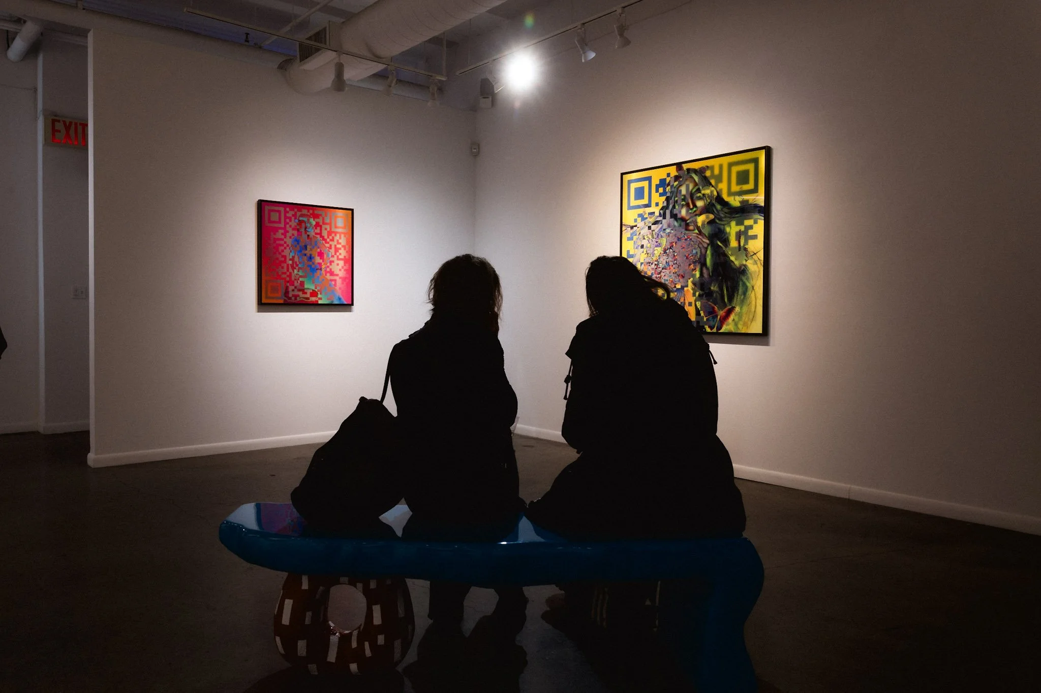 Deux personnes assises sur un banc dans une galerie d'art, regardant des œuvres colorées et abstraites accrochées à un mur.