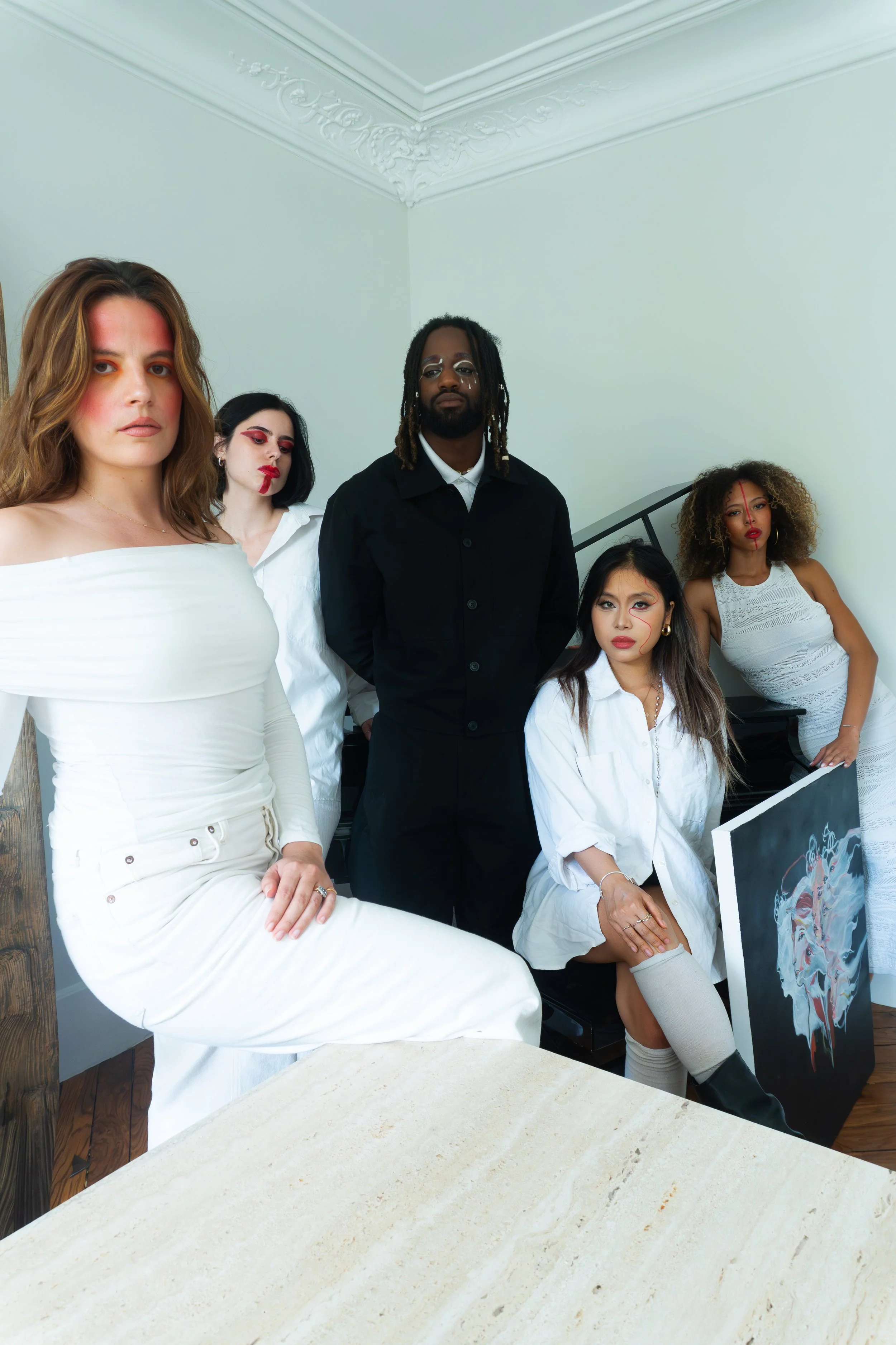 Un groupe de cinq personnes posant dans une pièce avec des murs blancs et une cheminée décorative. Chaque personne porte des vêtements blancs ou noirs avec du maquillage artistique sur le visage.