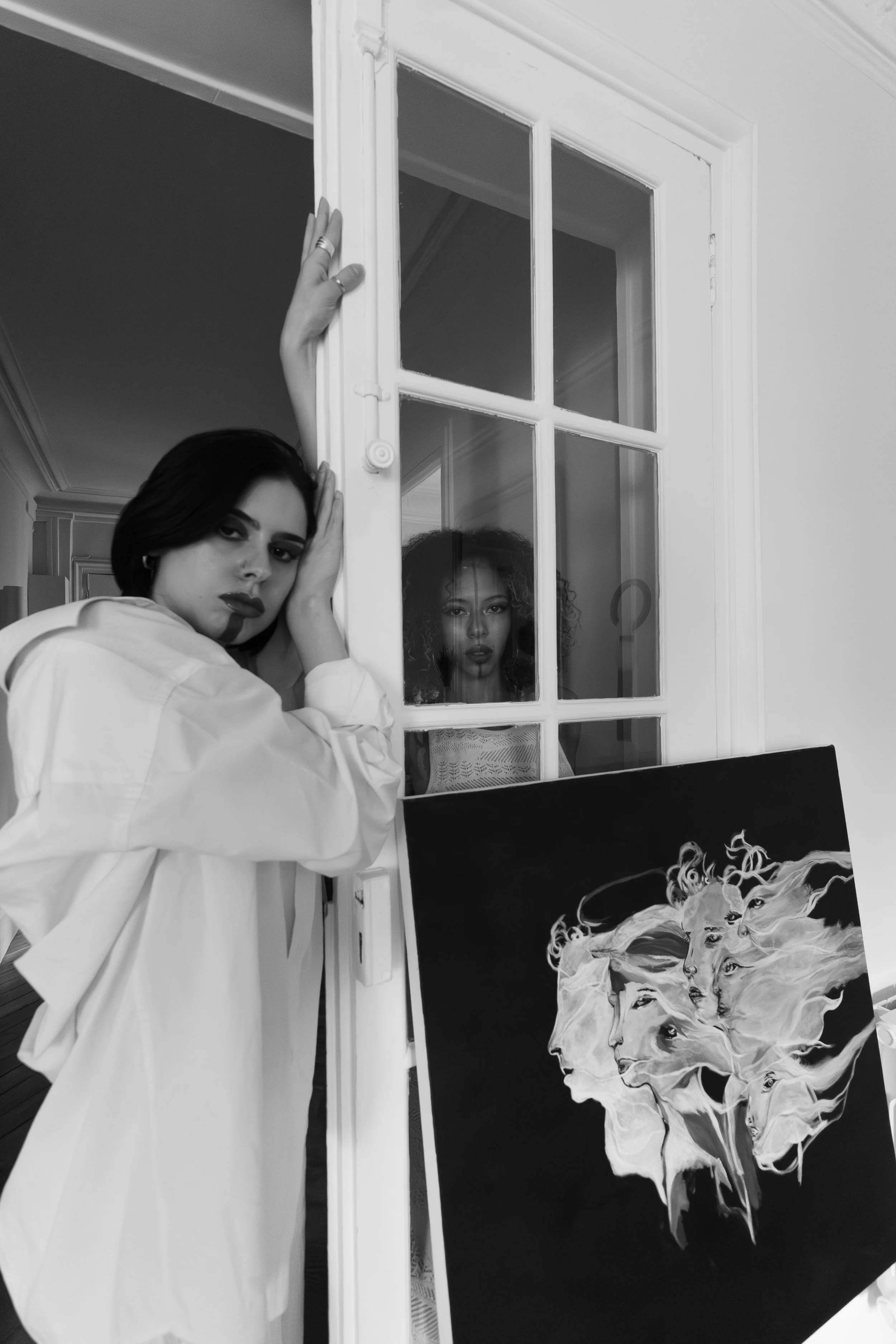 Deux femmes dans un espace intérieur, une en avant de la porte en train de poser pour la photo, une autre derrière la vitre de la porte. Il y a une peinture de visages variés à côté d'elles.
