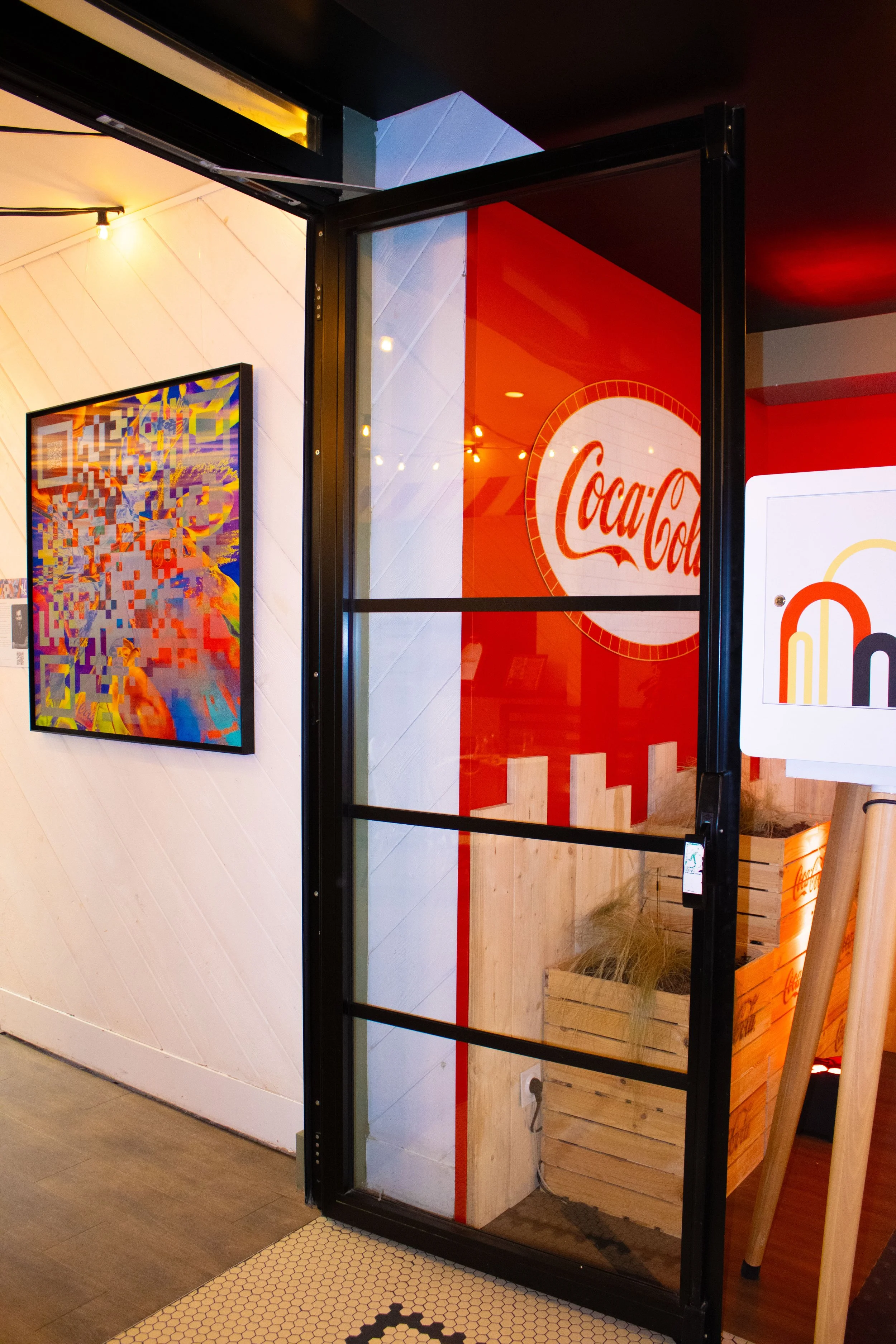 L'entrée d'un restaurant ou d'un café avec un mur rouge affichant le logo Coca-Cola et une porte en métal noir. À gauche, il y a une peinture colorée sur le mur blanc. La décoration intérieure est chaleureuse avec un éclairage tamisé.