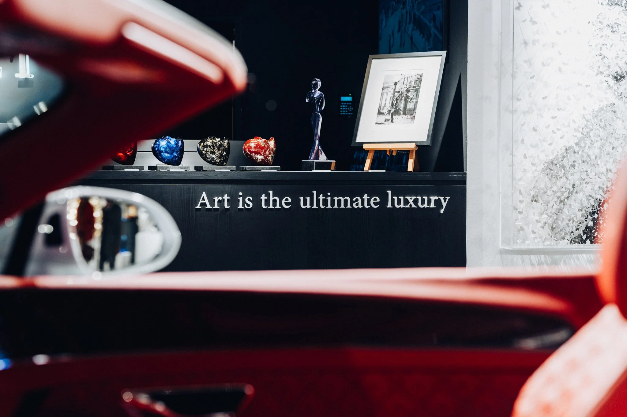 Une exposition d'art avec des sculptures, une peinture encadrée, et des cœurs en verre sur un piédestal noir portant la phrase "Art is the ultimate luxury" (L'art est le luxe ultime)