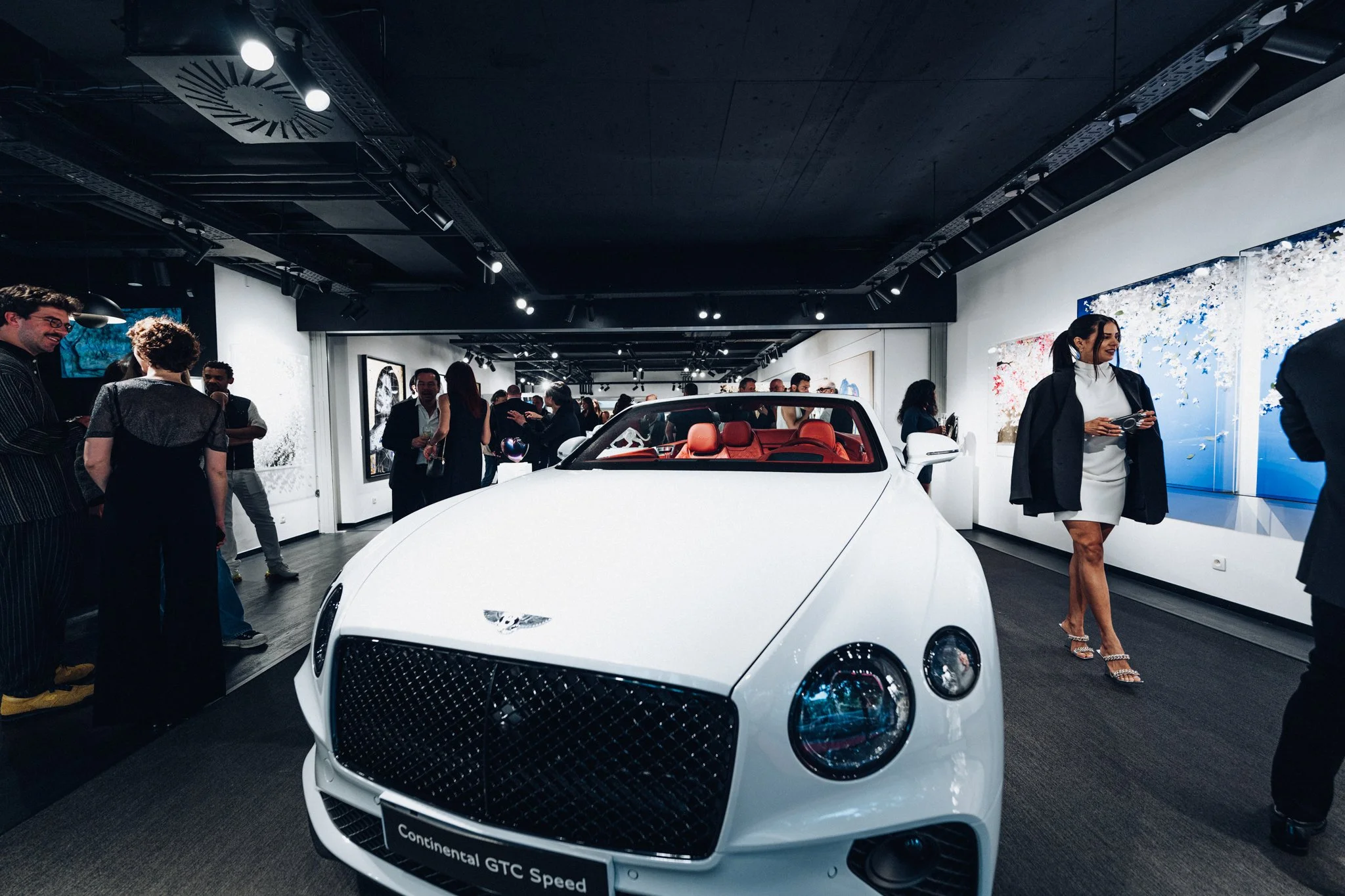 Voiture blanche de luxe exposée dans une galerie d'art lors d'un événement, avec des personnes regardant autour et des œuvres d'art sur les murs.