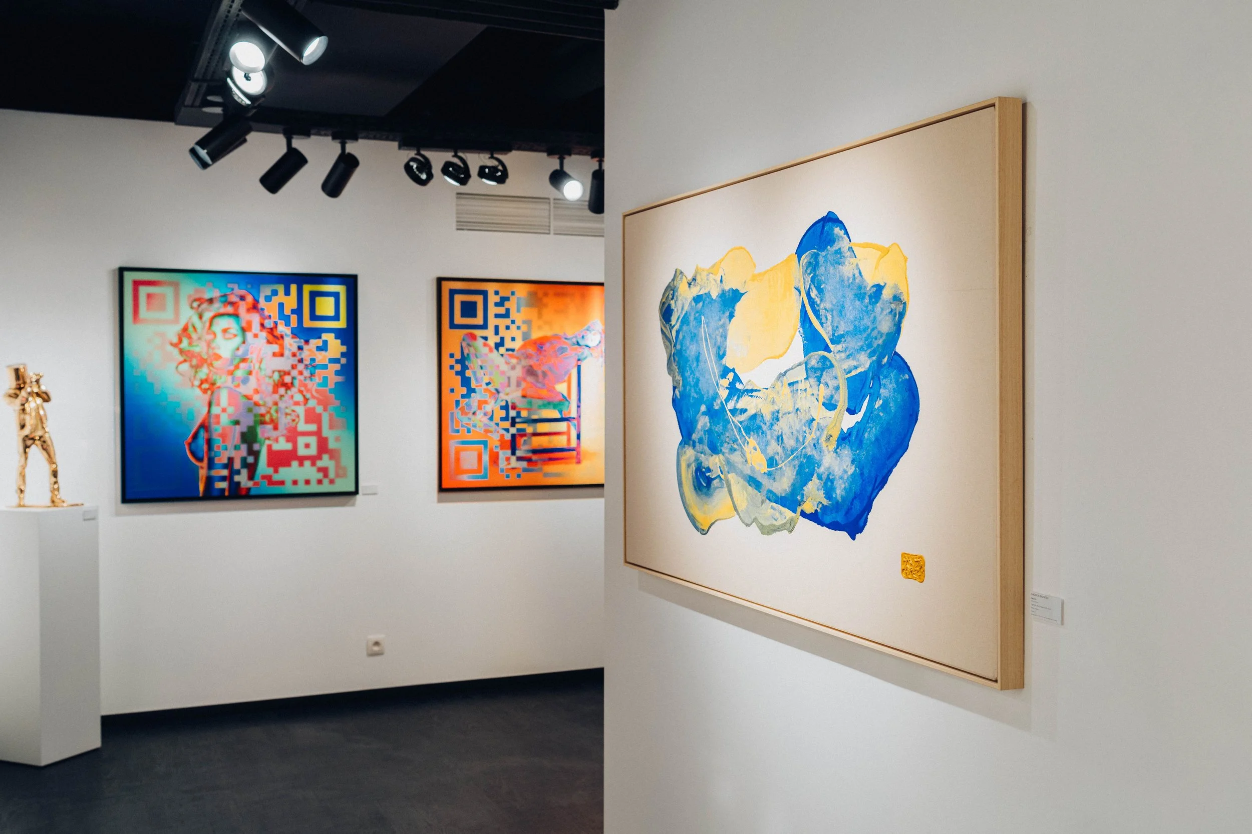 Exposition d'art contemporain dans une galerie, peinture abstraite avec des couleurs bleues, jaunes, et blanches sur un mur en bois, images numériques colorées à l'arrière-plan.