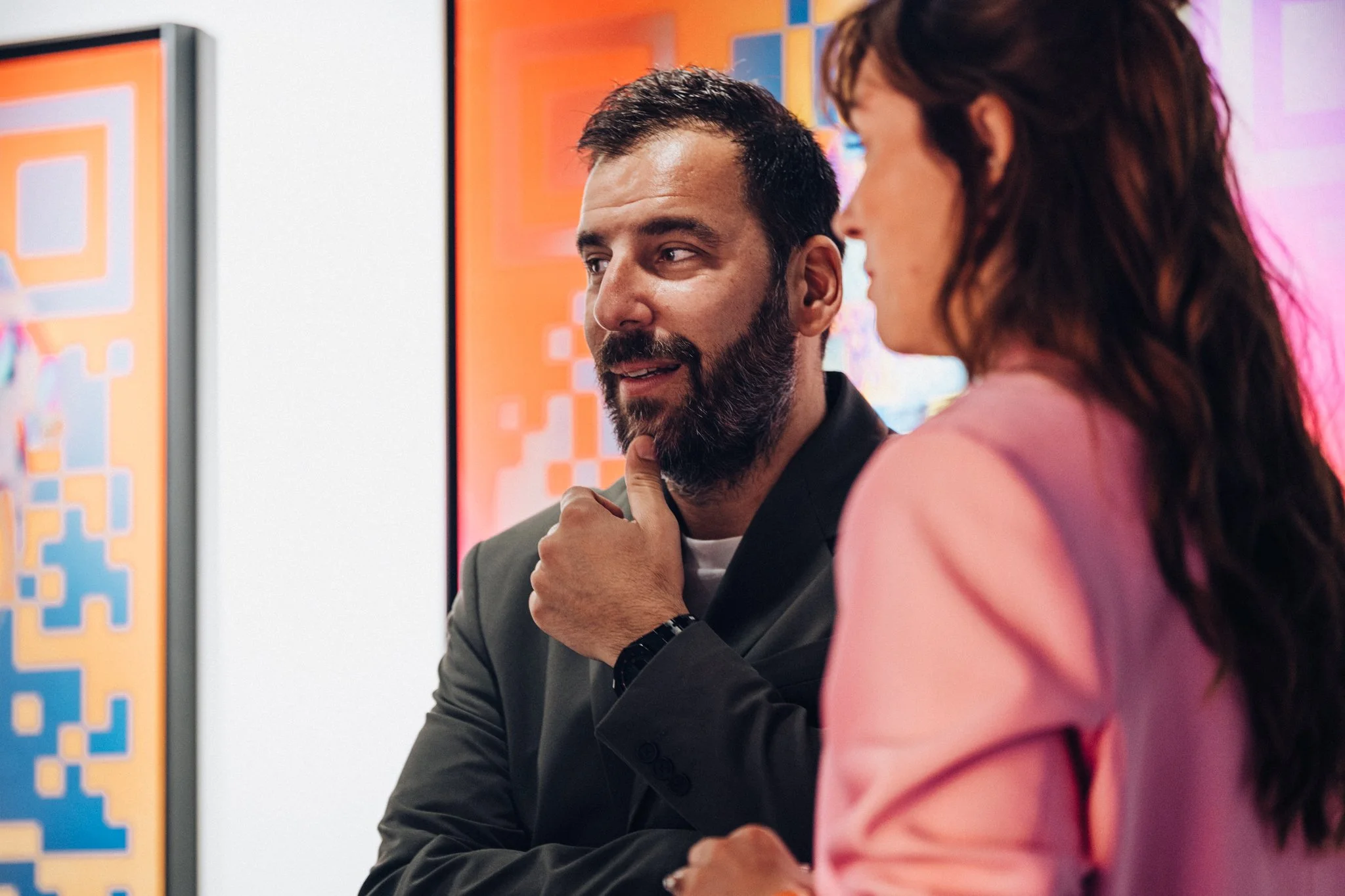 Deux personnes discutent, un homme avec une barbe et une femme avec des cheveux longs, dans un environnement coloré et moderne.