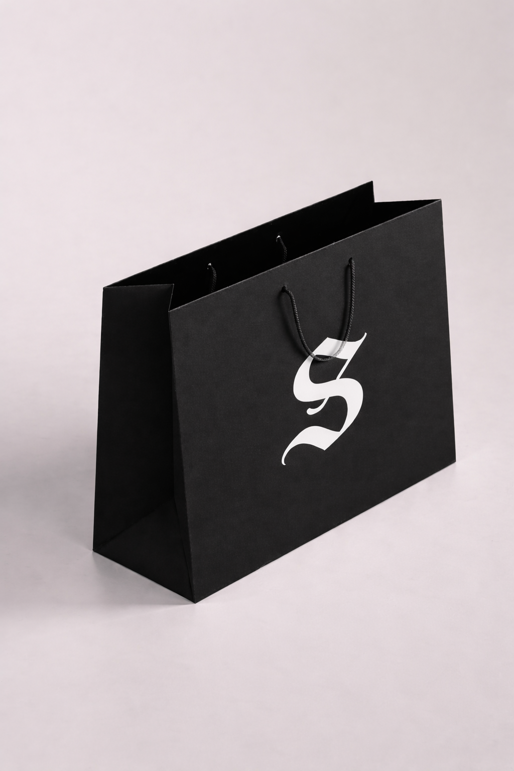 Sac en papier noir avec une poignée en corde noire et un logo blanc stylisé en forme de S ou de serpent.