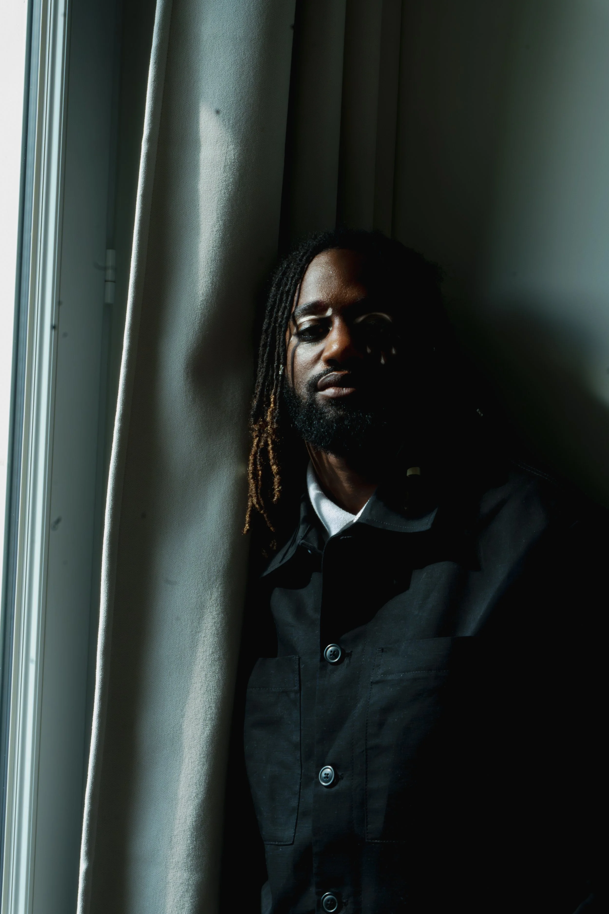 Homme noir aux dreadlocks, portant une veste noire, se tenant près d'une fenêtre avec rideaux, dans un intérieur sombre.