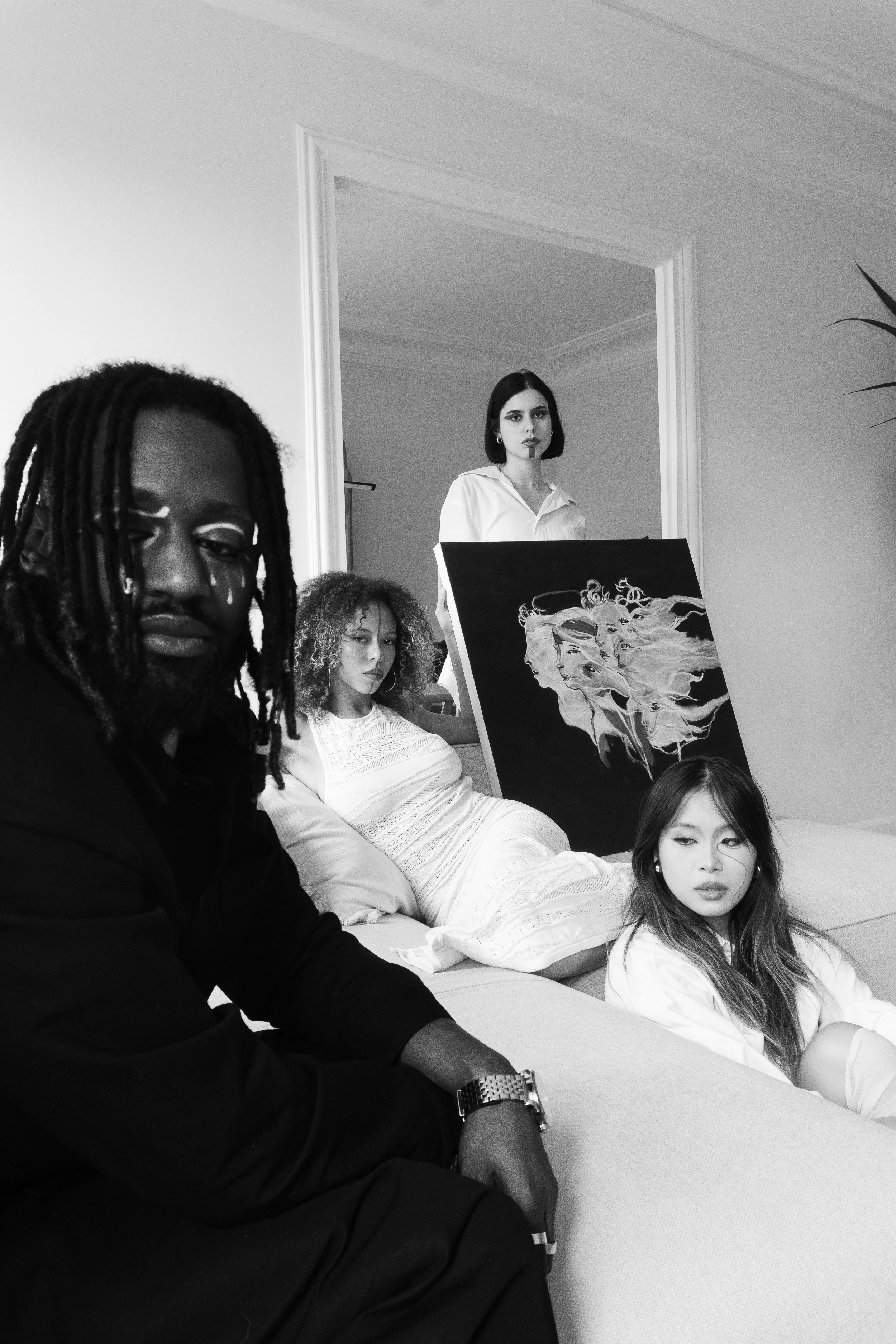 Groupe de quatre personnes dans une pièce, deux femmes assises sur un canapé, une femme debout derrière une peinture, et un homme en avant-plan à gauche, dans un style artistique noir et blanc.