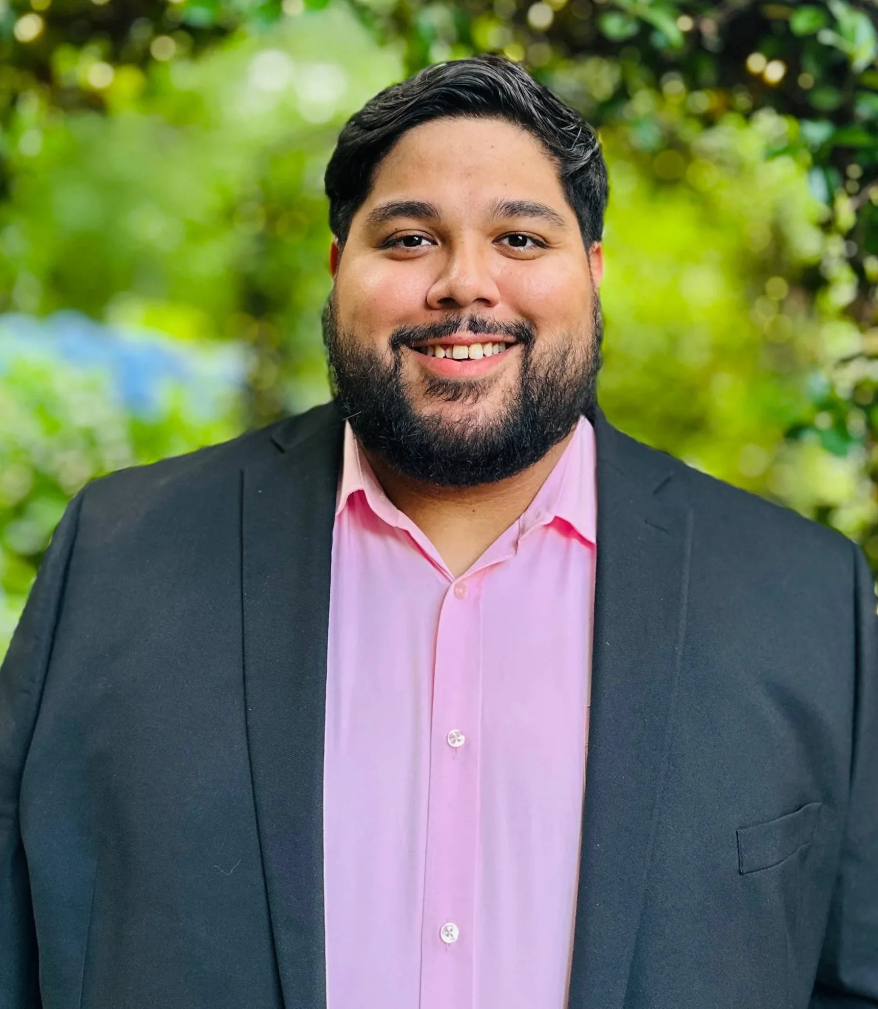 A man with dark hair and a beard, wearing a pink shirt and black blazer, smiling outdoors with a blurred green background.