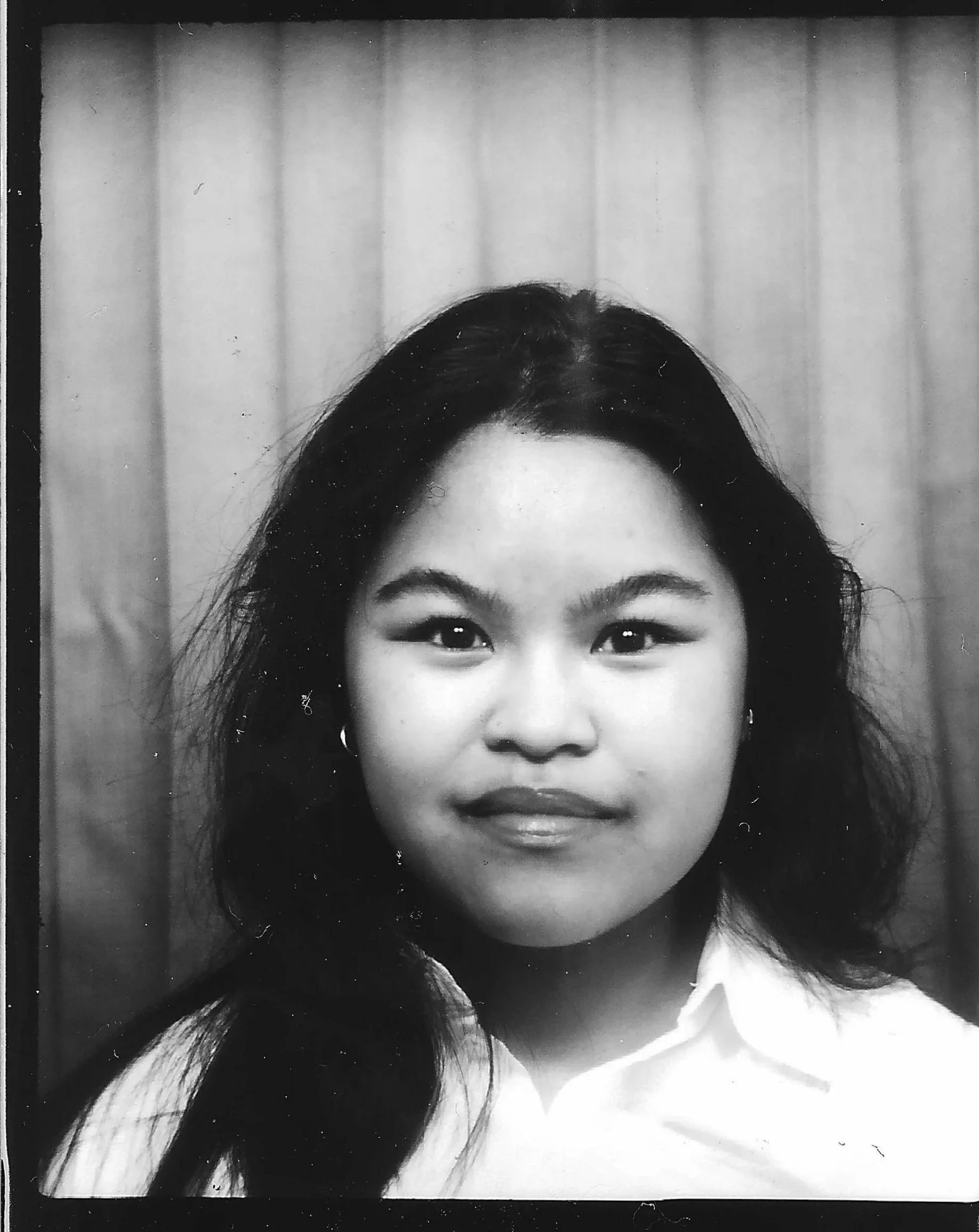 Black and white portrait of a young woman with dark hair, wearing earrings and a white shirt, standing in front of a wooden-paneled wall.