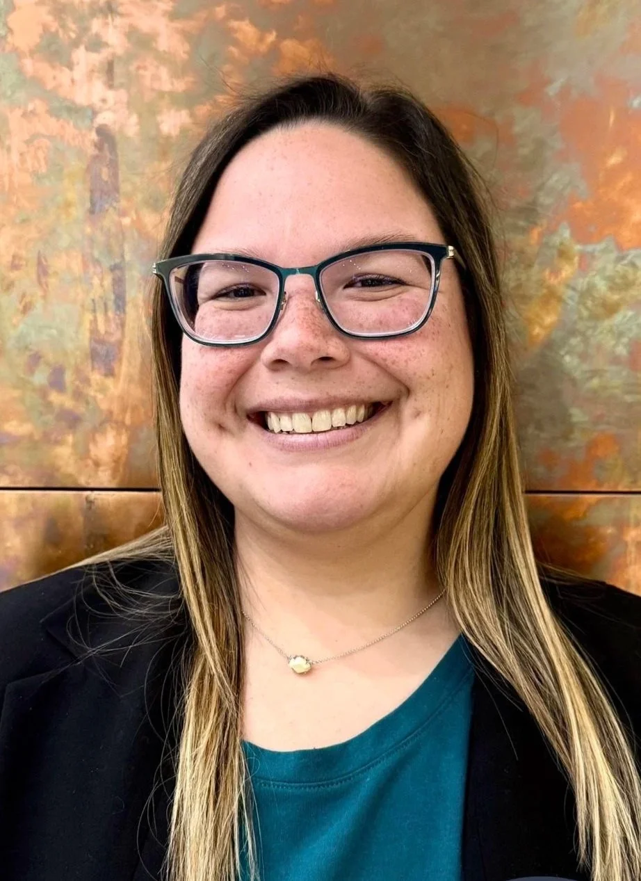 A woman smiling, wearing glasses, a black blazer, and a teal shirt, standing in front of a colorful, abstract background.