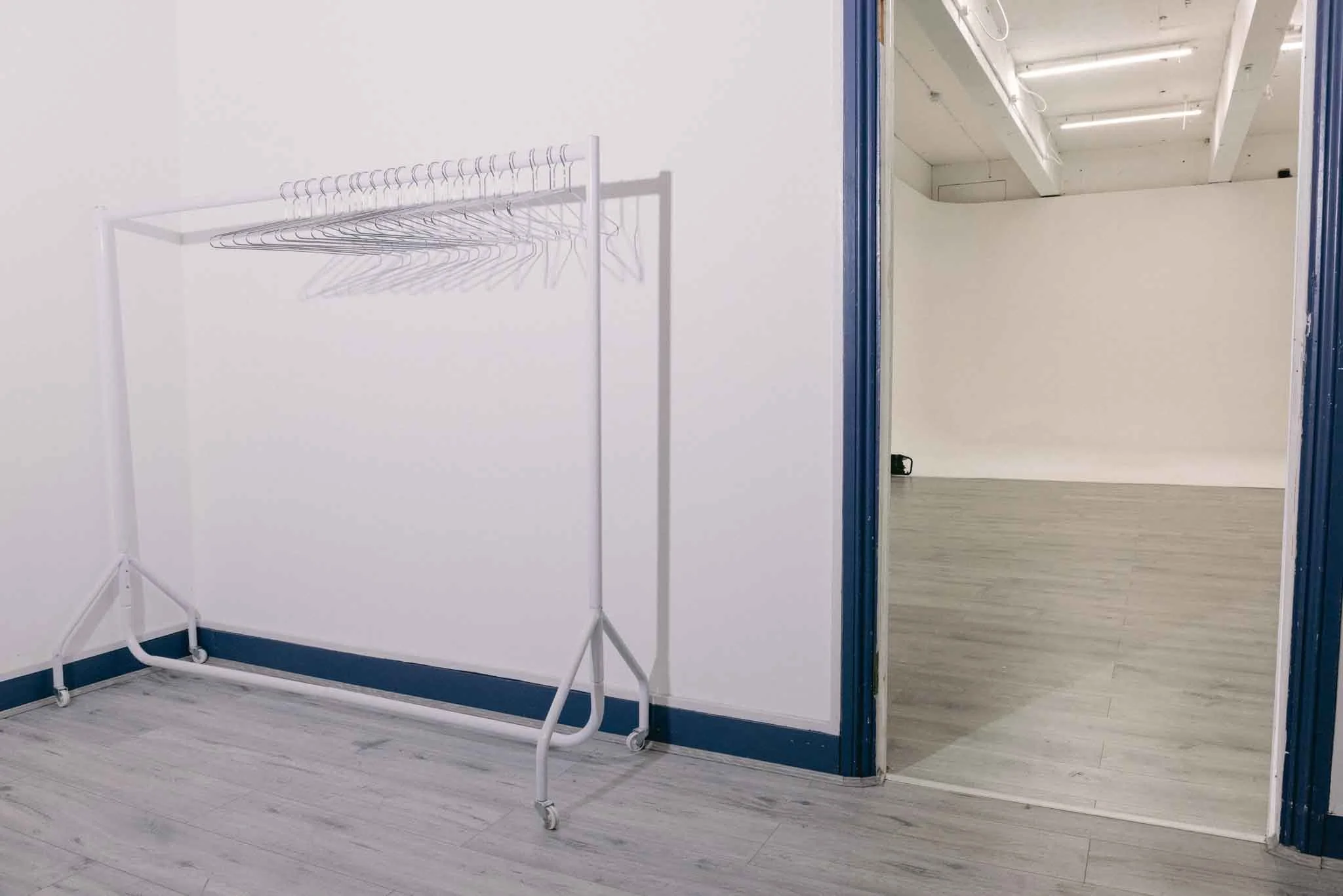 Empty room with white walls and light wooden flooring. A white metal clothing rack with wheels is positioned against one wall. An open doorway reveals another room with a ceiling of exposed beams and pipes, painted white, and identical light wooden flooring.