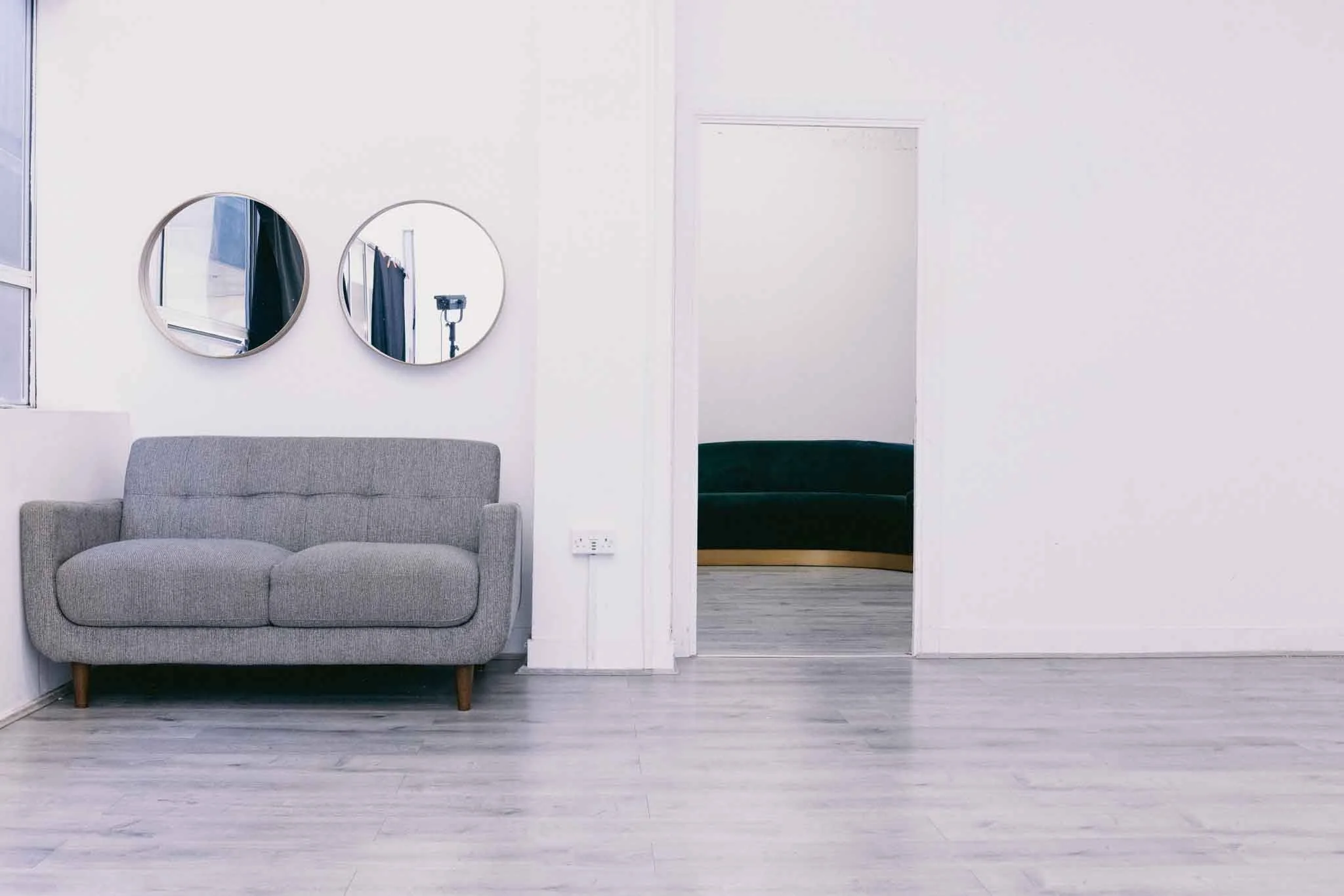 A gray fabric sofa with wooden legs in a minimalistic white room. Two round mirrors are mounted on the wall above the sofa. An open doorway reveals a dark green couch inside another room.