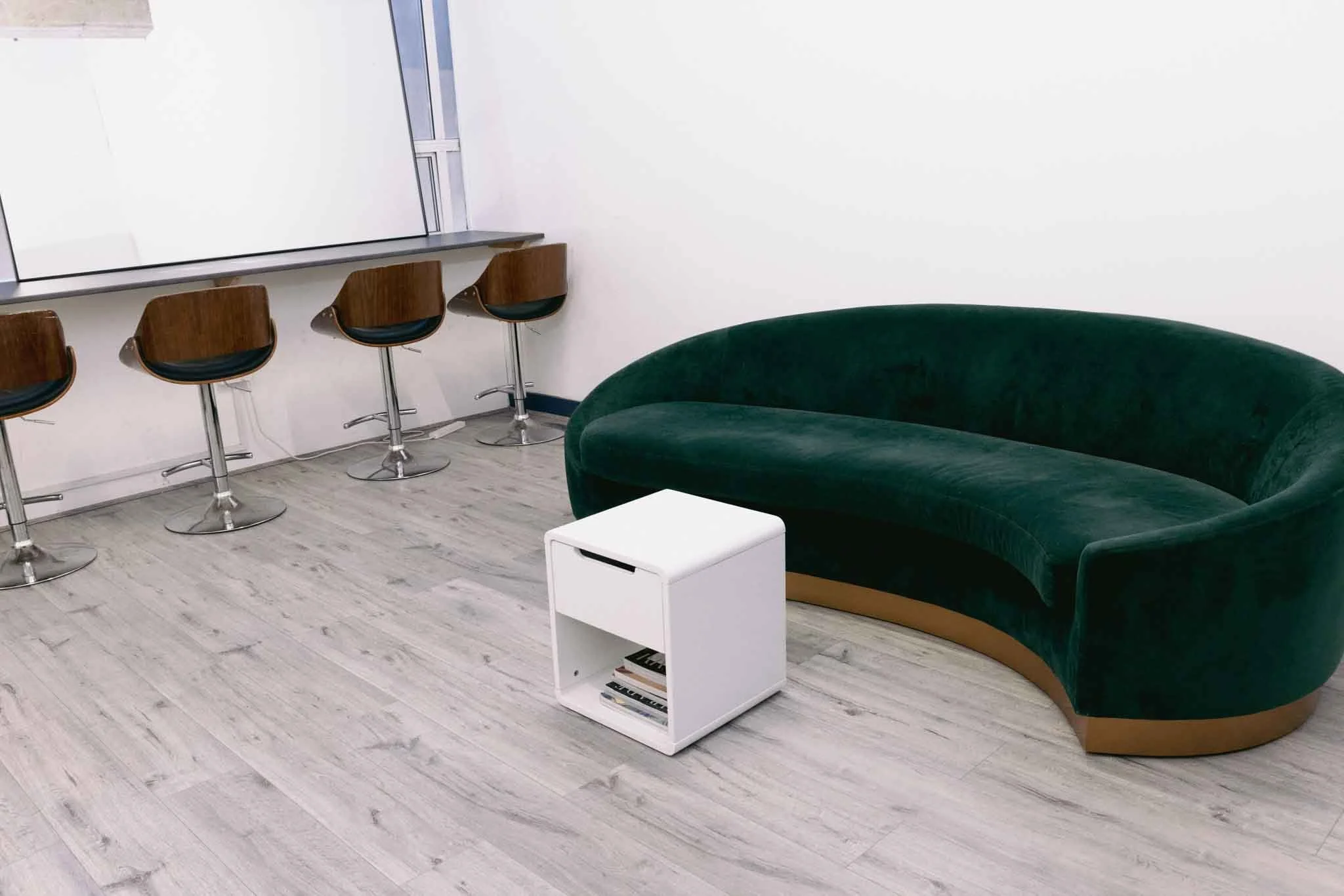 A modern lounge with a green velvet curved sofa, a white small side table with books underneath, a white wall, and a row of three wooden barstools with black seats at a high counter.