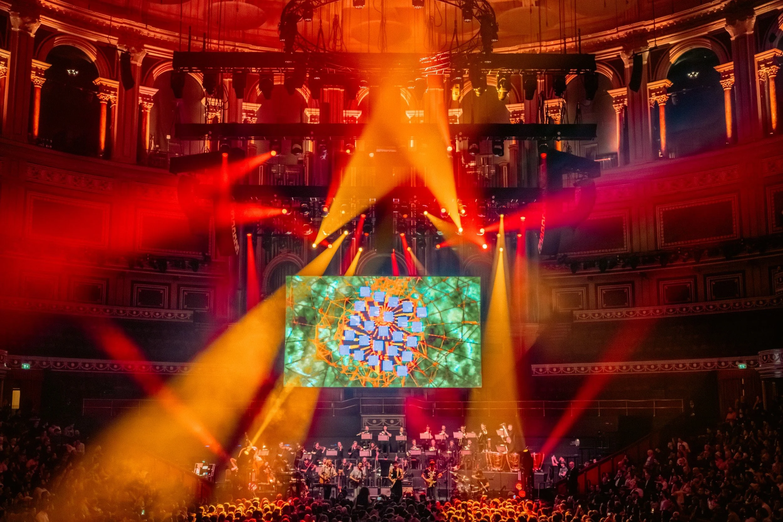 Concert hall with red and yellow stage lighting, large digital screen displaying colorful abstract art, and a band performing.