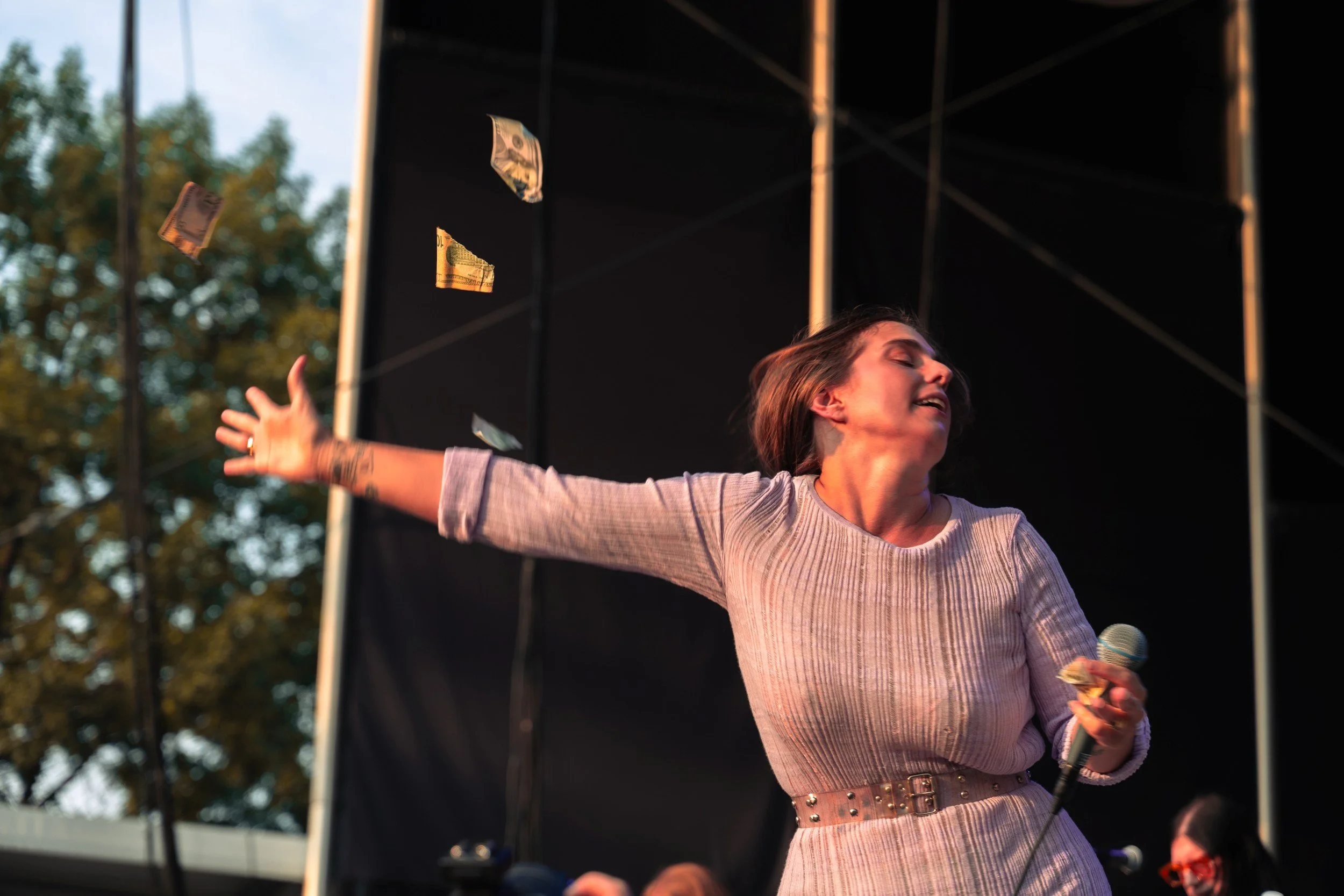 A woman with brown hair and light skin, wearing a light-colored ribbed top and a wide belt, is on stage at an outdoor event during sunset, holding a microphone in one hand and gesturing with her other hand while money falls from above.