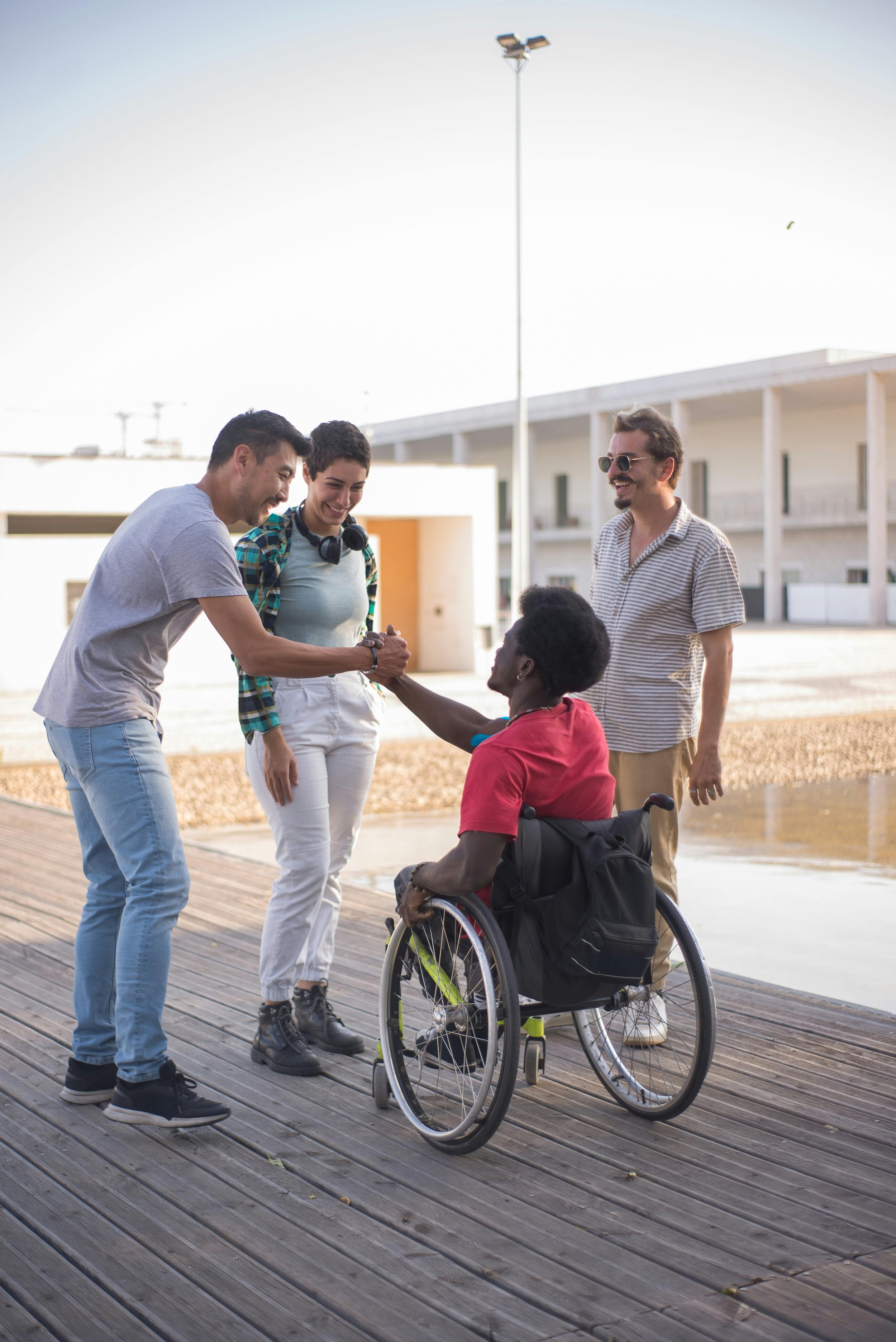 Two men and a woman are shaking hands with a woman in a wheelchair outdoors near a body of water, with a modern building in the background.