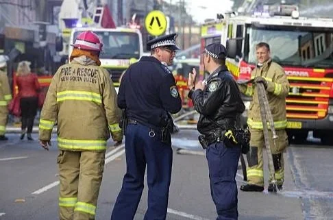 Firefighters and police officers talking on a street with emergency vehicles in the background.