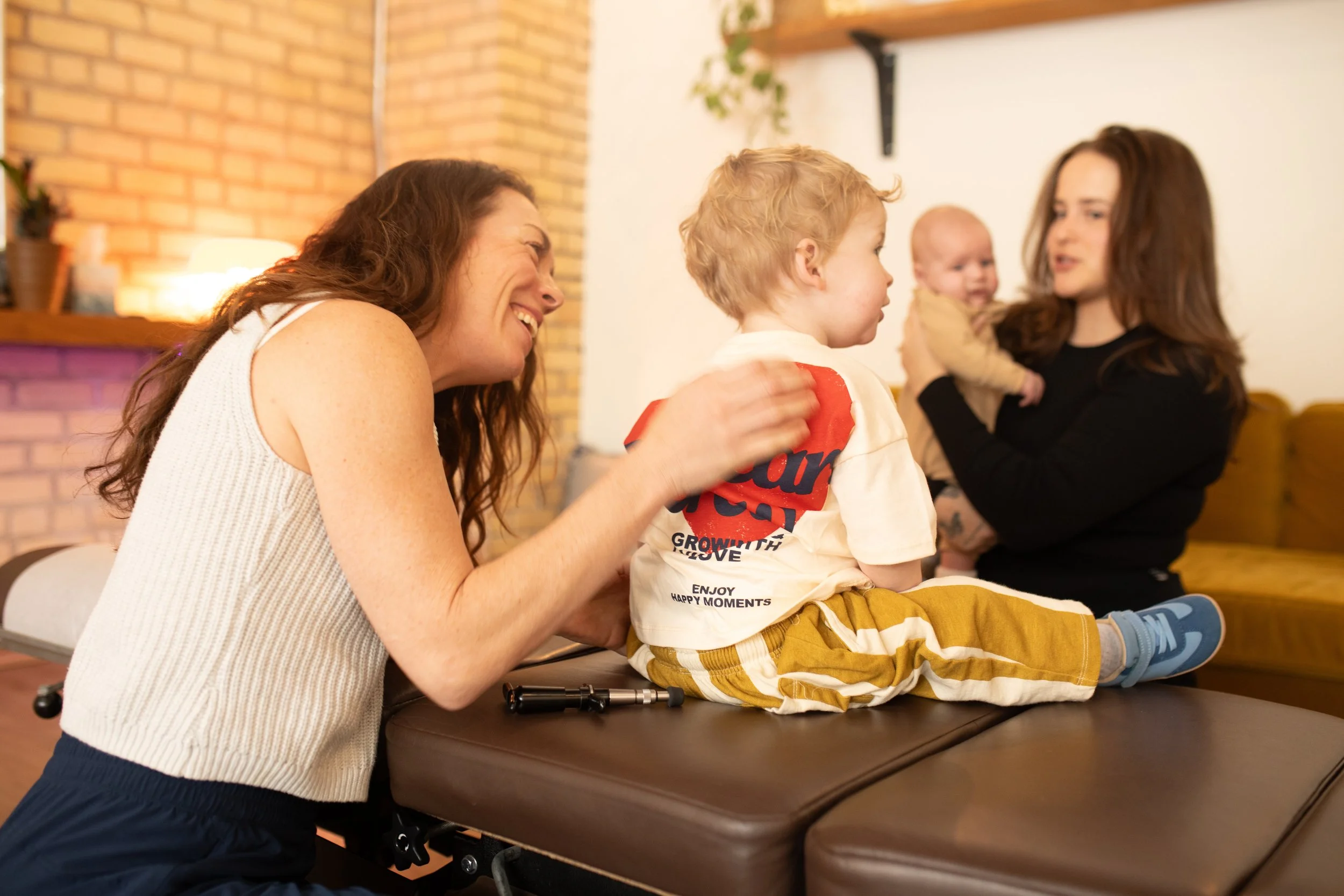 A woman laughs as she helps a young boy sit on an examination table, while another woman holding a baby looks on in a cozy room with a brick wall and warm lighting. Baby receiving a chiropractic adjustment