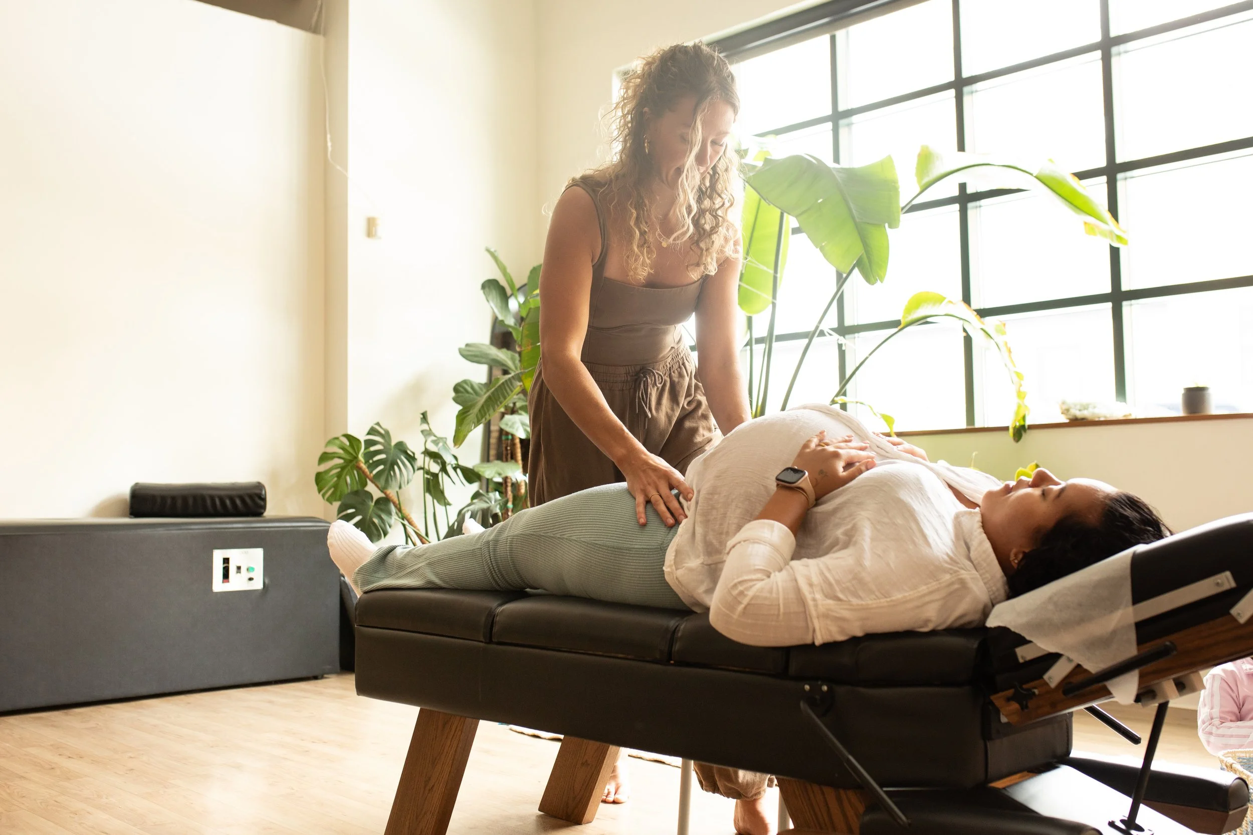 A woman receiving a massage on her abdominal area from a female massage therapist in a room with large windows and green plants.
