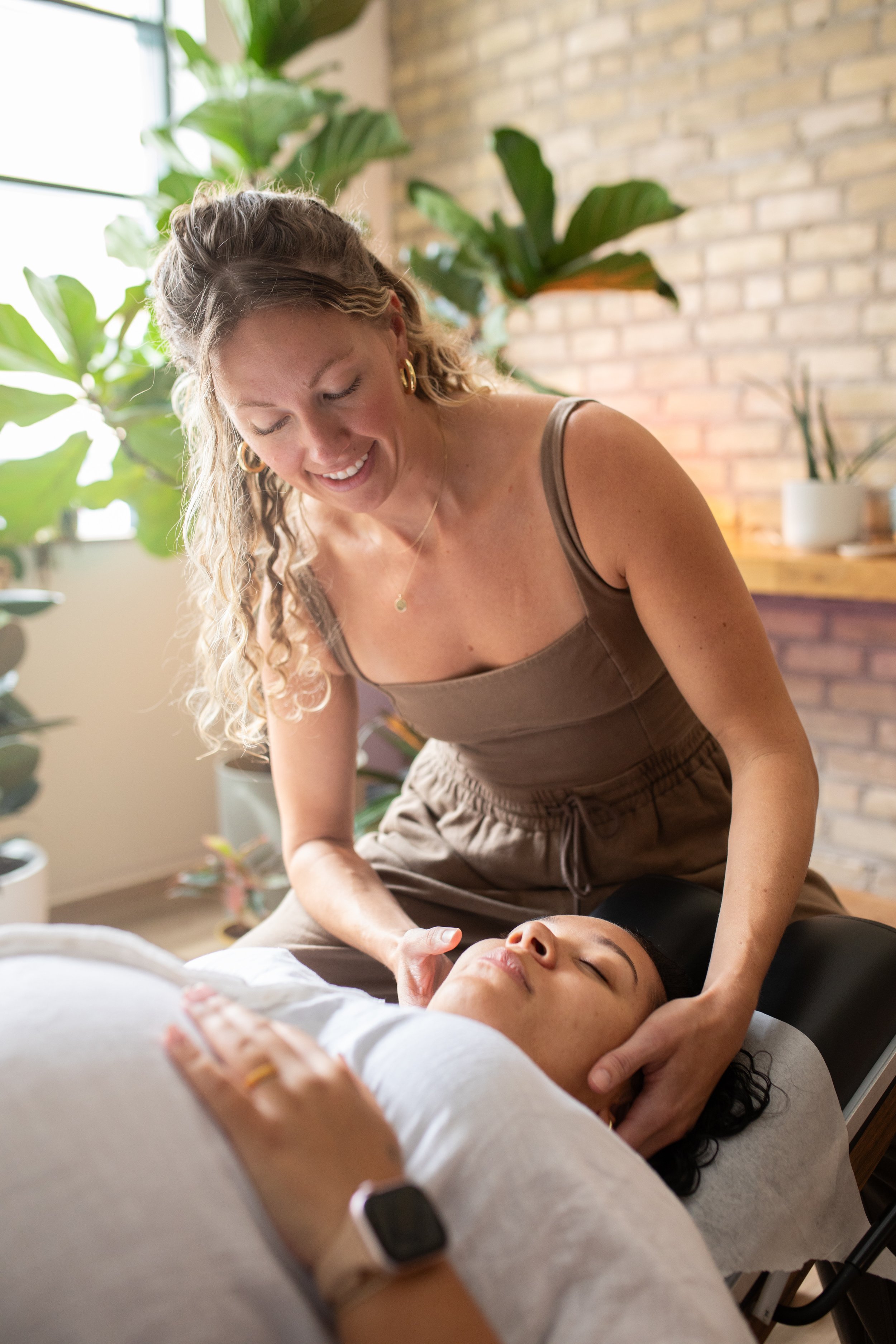 A woman giving a facial massage to a woman lying on a treatment bed in a spa-like setting with green plants and a brick wall in the background.