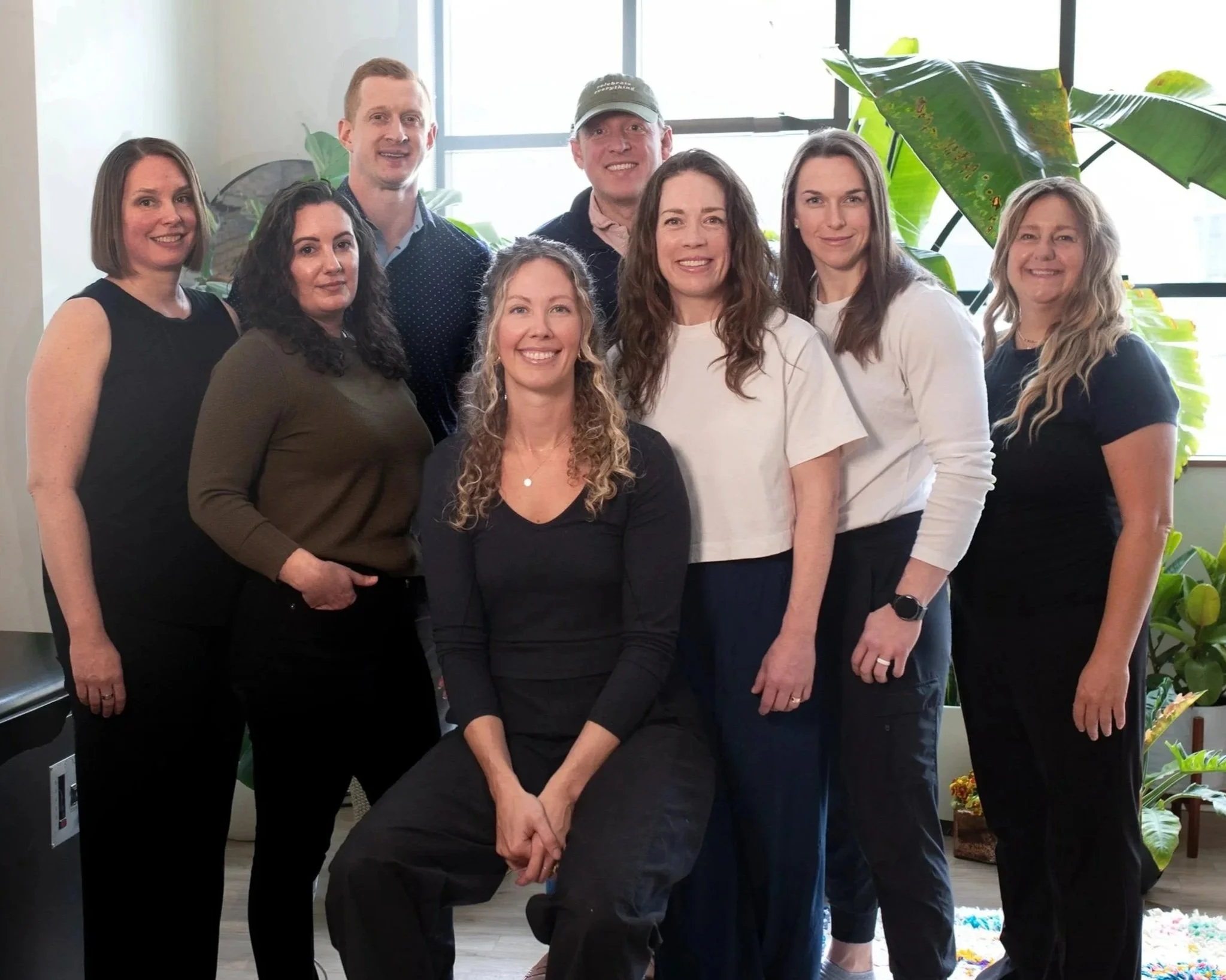 A group of ten people, mostly women, standing and sitting in a bright indoor space with large green leafy plants and a window in the background.