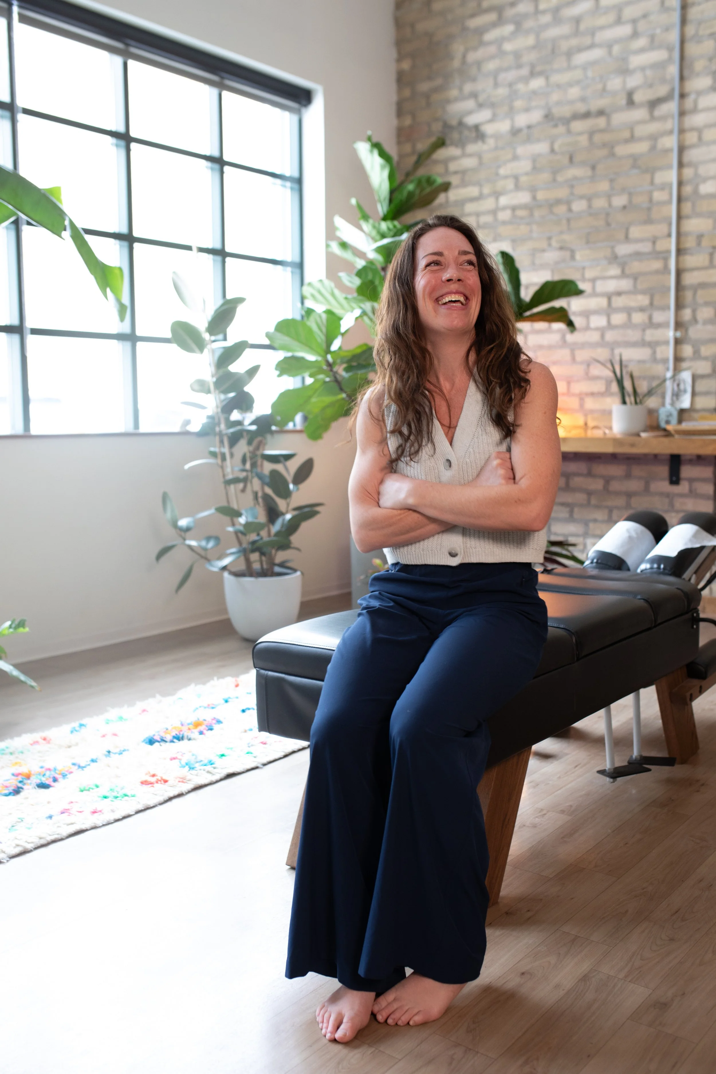A woman sitting on a massage table in a bright, modern room with large windows, plants, and a brick wall, smiling and laughing.