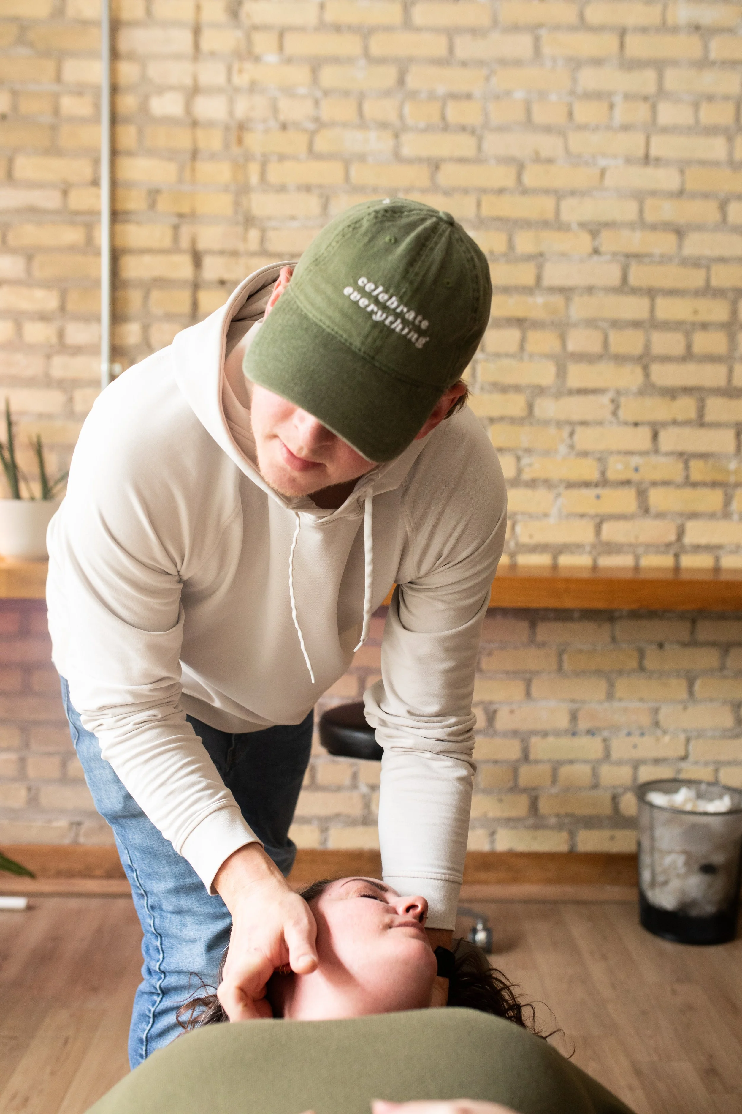 A person giving a neck massage to another person lying down, in a room with a brick wall background.