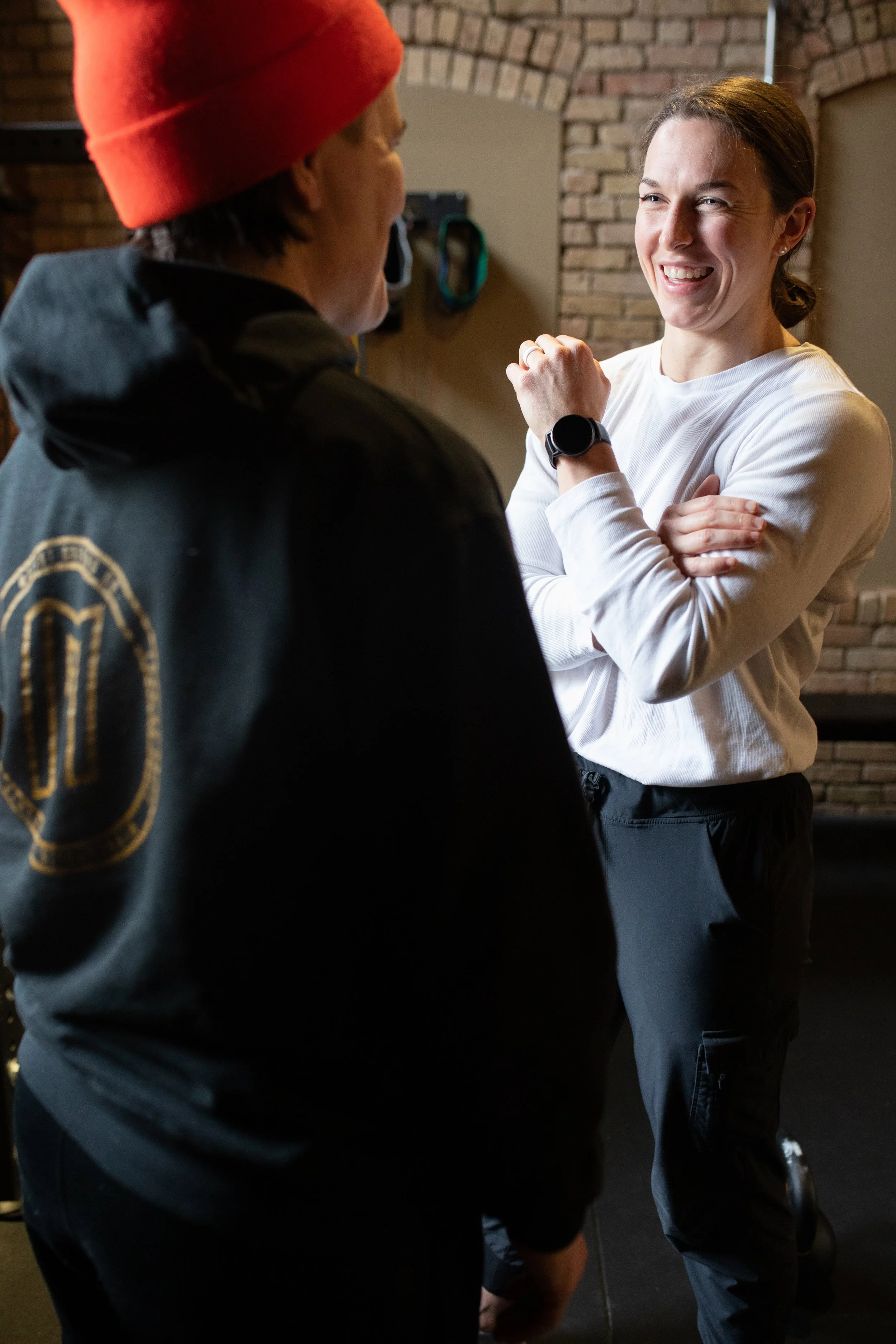 Two women having a conversation indoors, one in a black jacket and red beanie, the other in a white long-sleeve shirt with crossed arms, smiling.