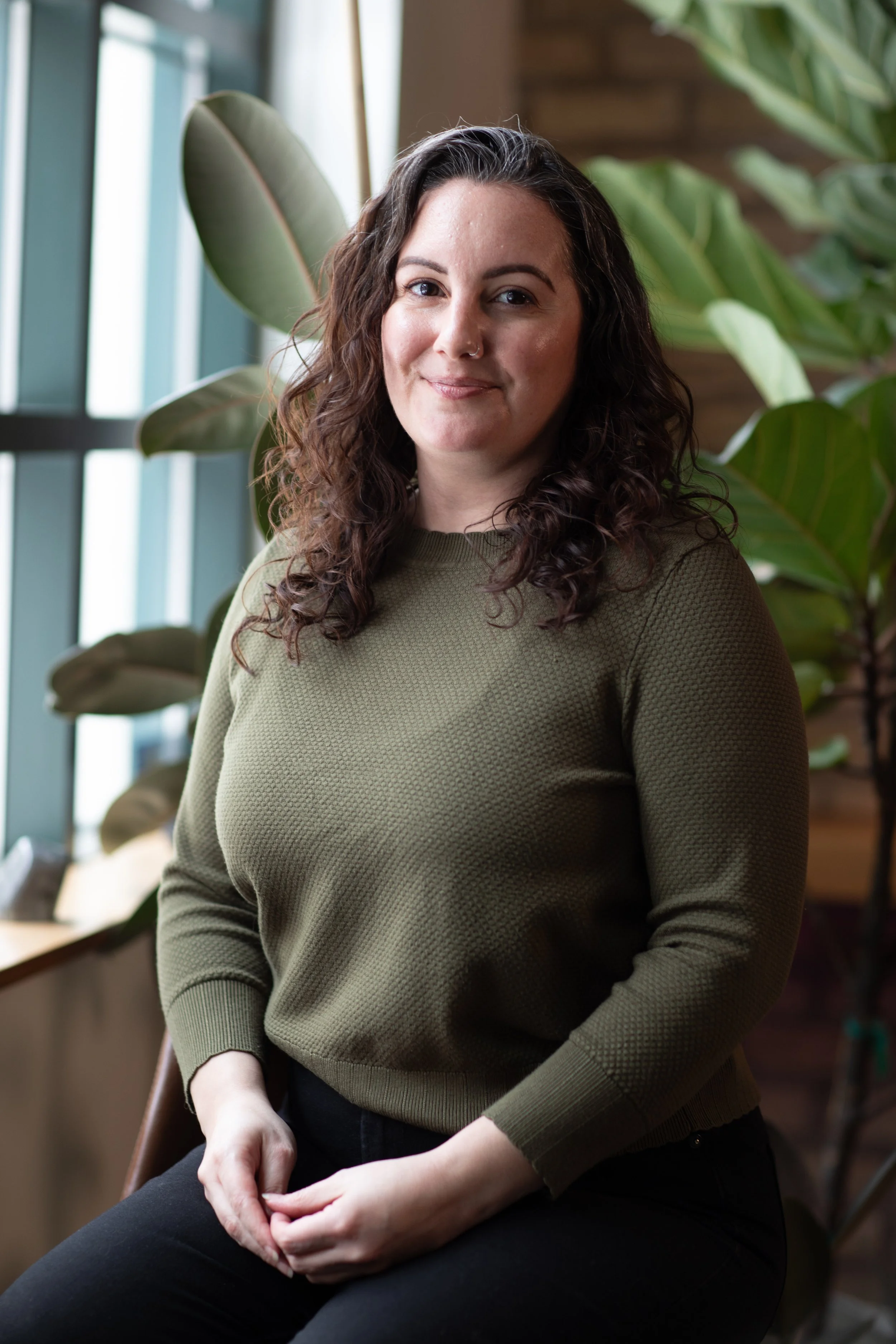 A woman with curly brown hair and light skin sitting indoors near large green houseplants, wearing an olive green sweater and black pants, smiling softly at the camera.