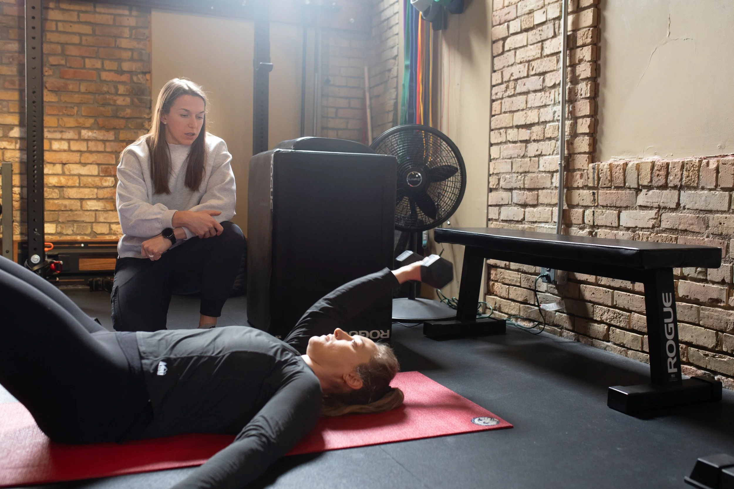 Two women in a gym, one woman lying on a red yoga mat and lifting a dumbbell, and another woman kneeling beside her observing.