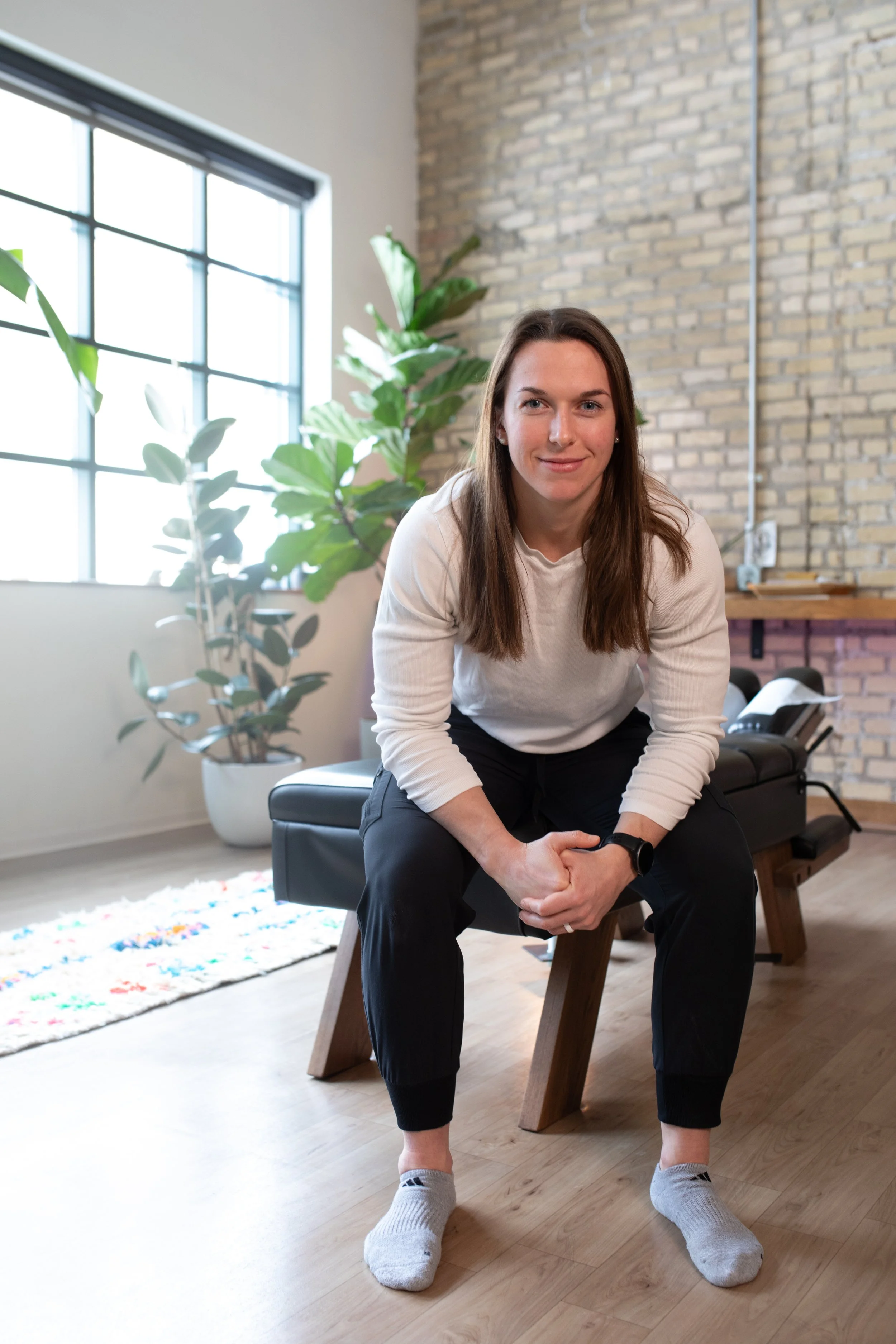 A young woman with long brown hair, wearing a white sweatshirt, black pants, and gray socks, sitting on a black wooden bench in a modern room with brick wall and large window, smiling at the camera.