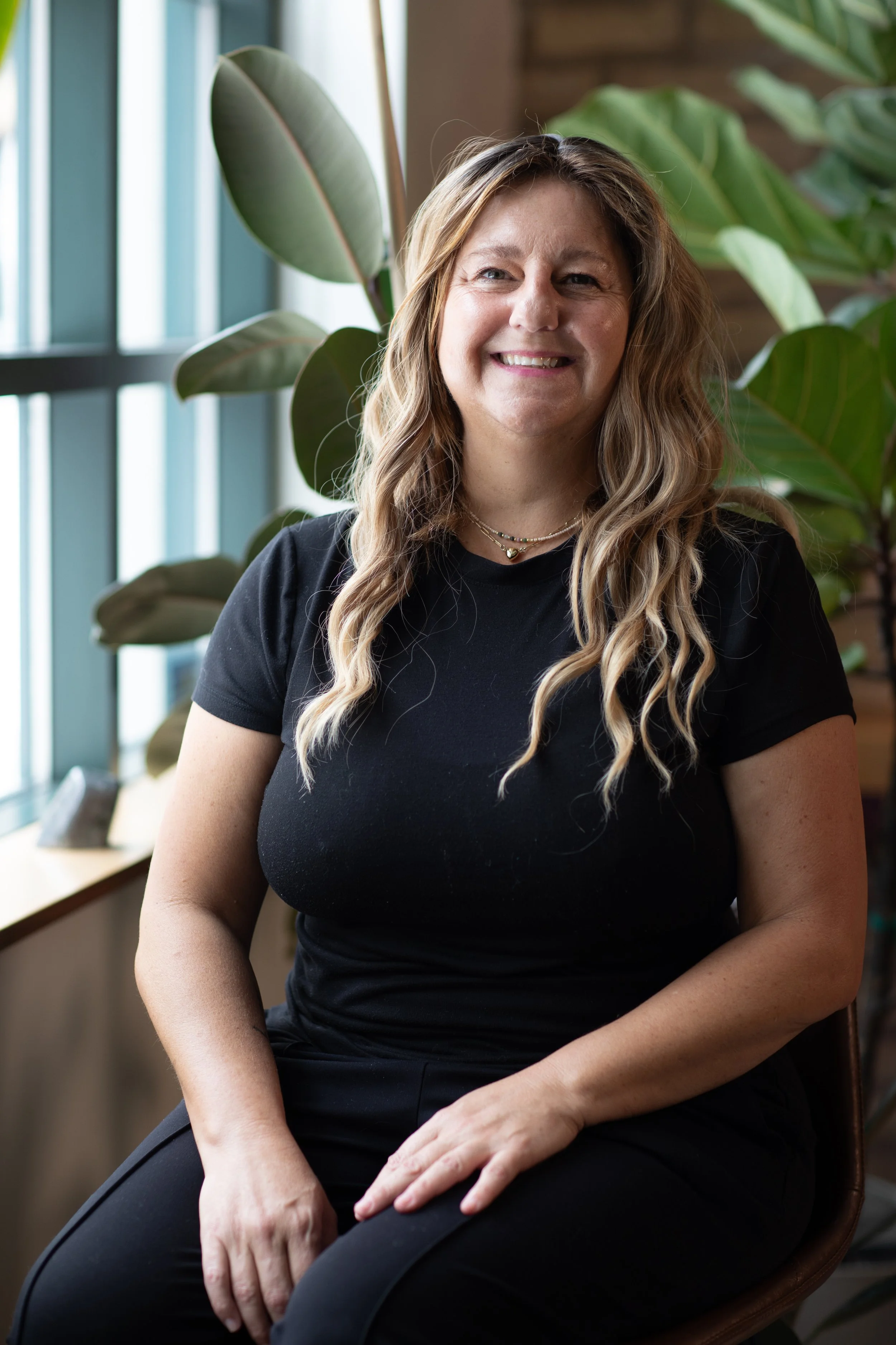 A woman with long blonde hair smiling, wearing a black t-shirt and necklaces, sitting in front of large green leaves and a window.